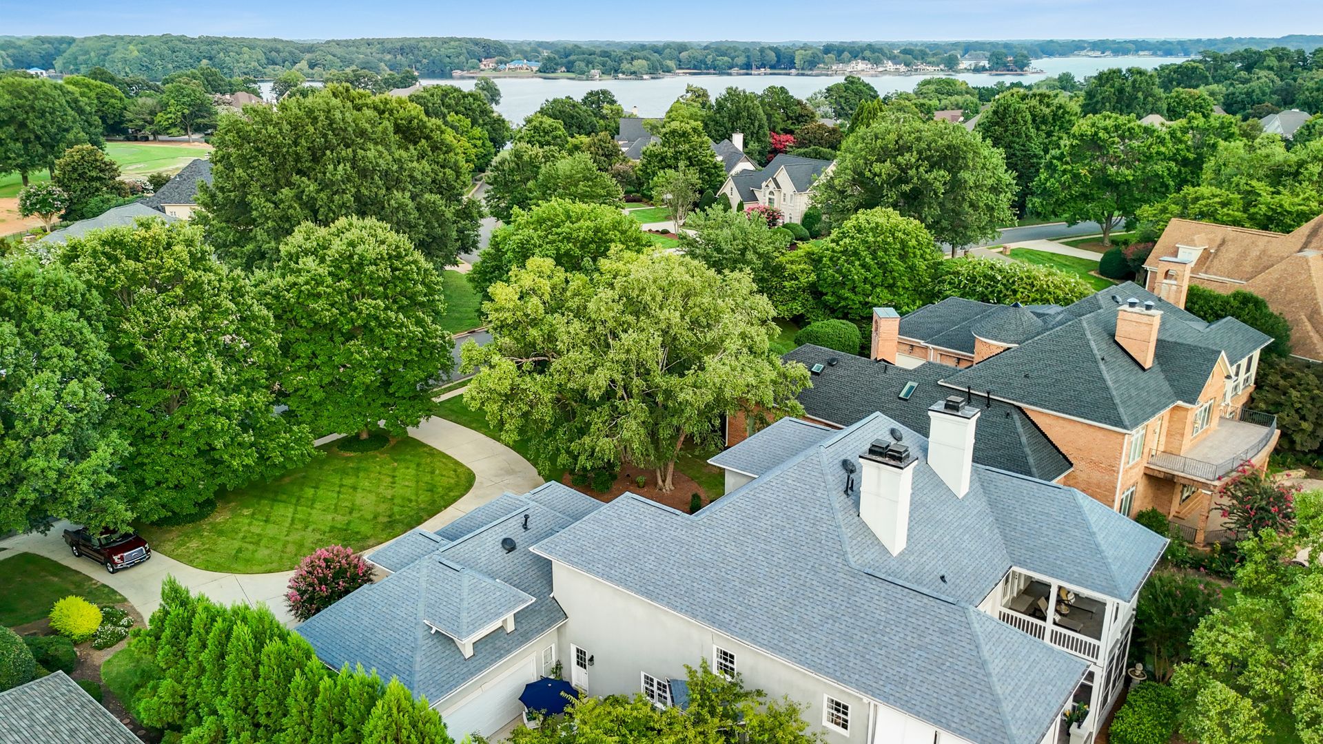 Aerial view of houses with gray roofs surrounded by green trees, near a lake.
