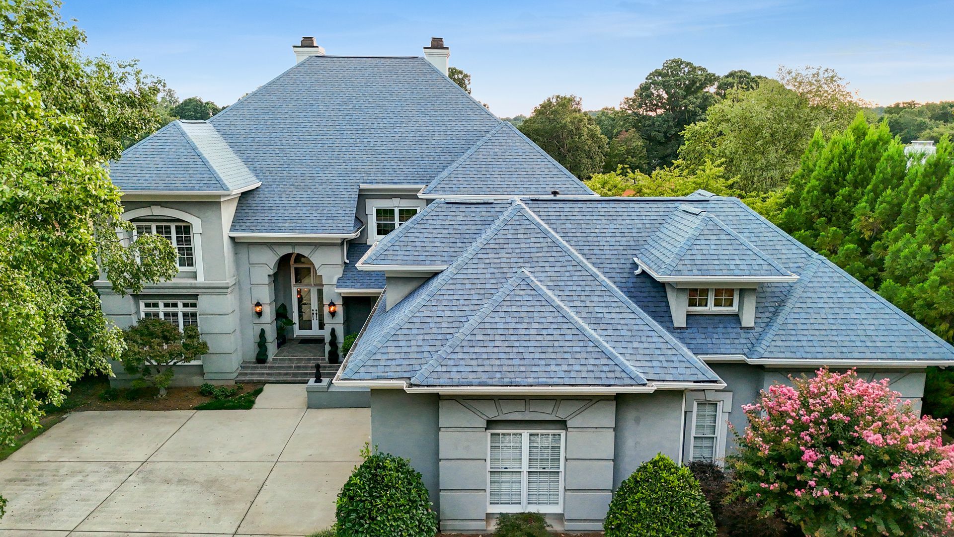 Gray stucco house with blue-gray tile roof, surrounded by green trees and pink flowering bush.