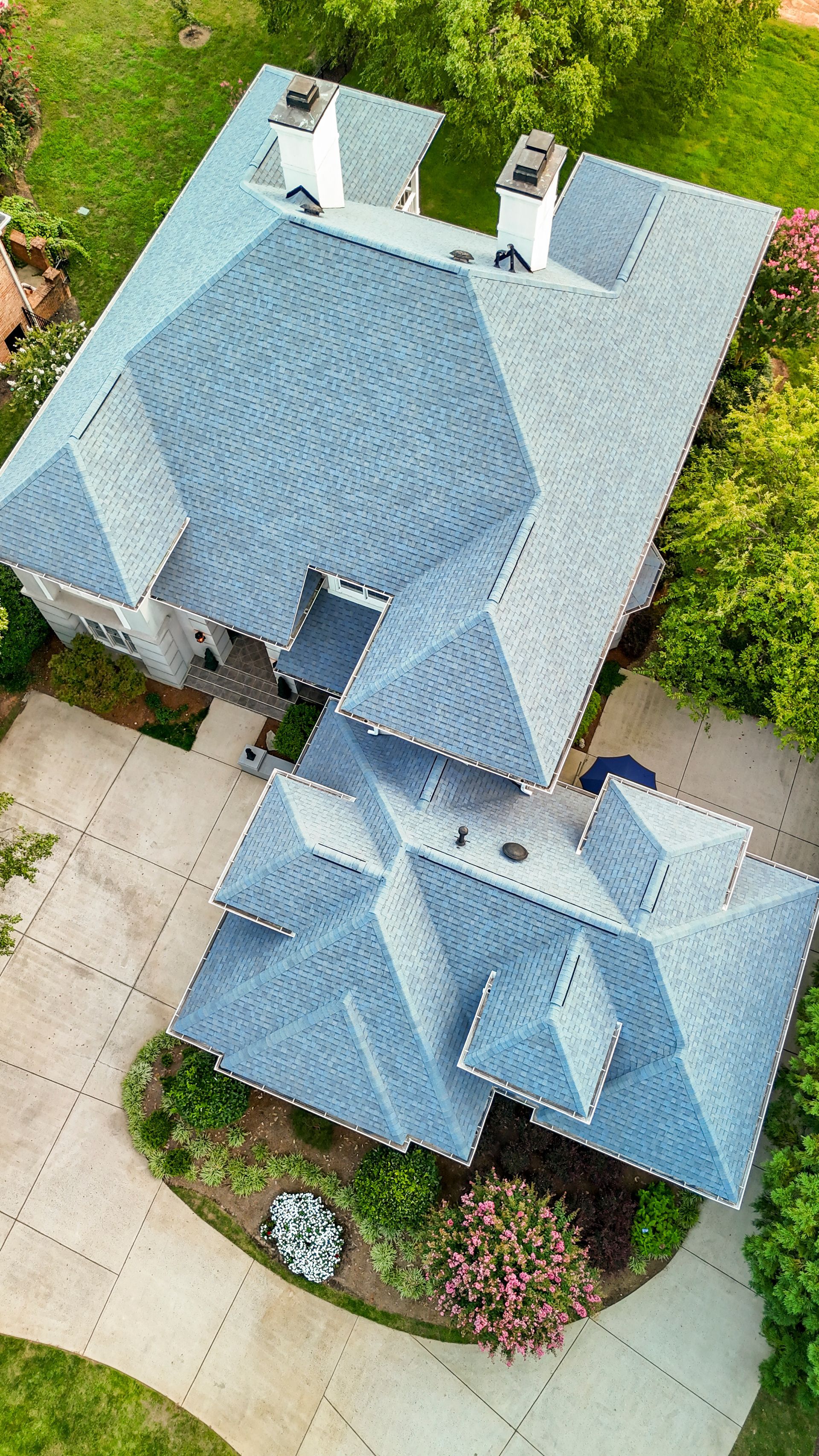 Overhead view of a house with a gray roof, two chimneys, a driveway, and surrounding greenery.