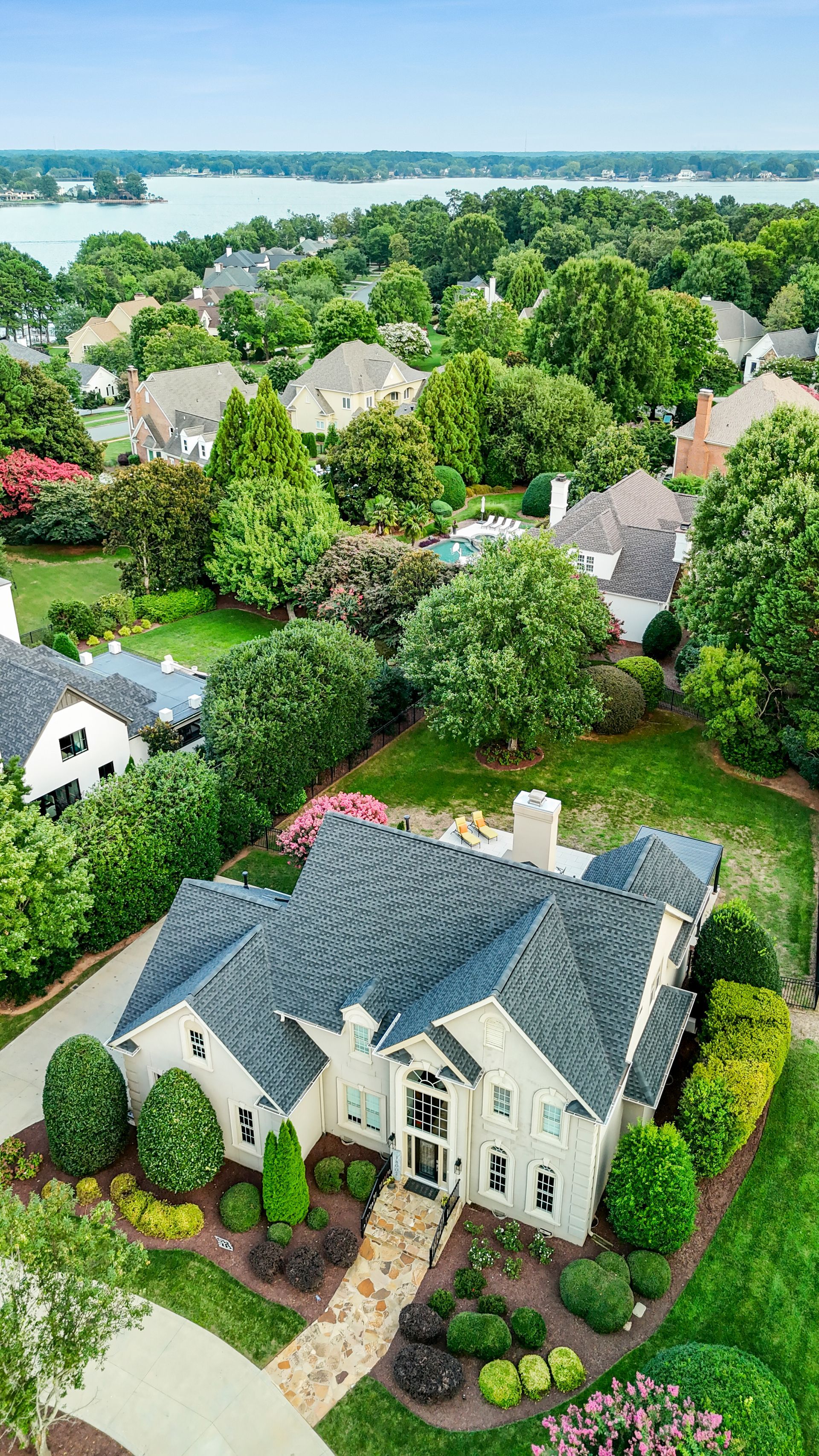 Aerial view of a large house with a dark gray roof, surrounded by green trees and other houses. A lake is in the background.