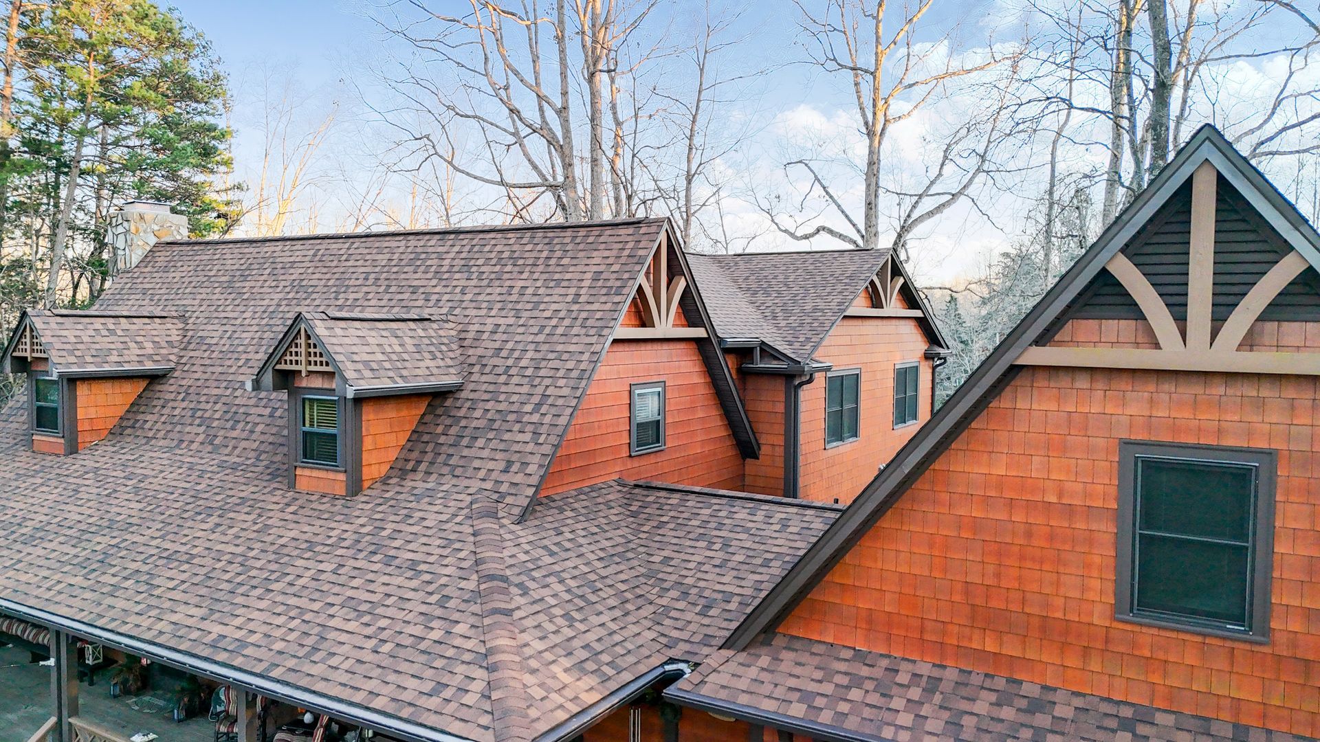 Brown shingled roof of a house with orange siding, gables, and dark trim against a backdrop of trees.