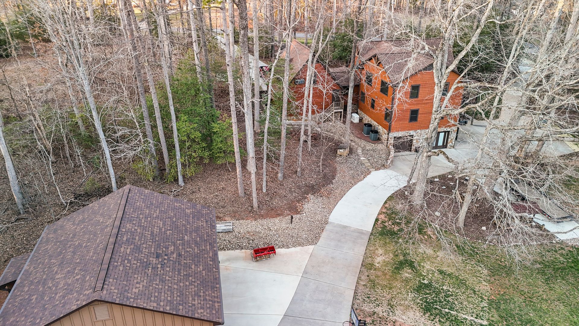 An aerial view of a brown wooden house and driveway surrounded by trees with bare leaves.