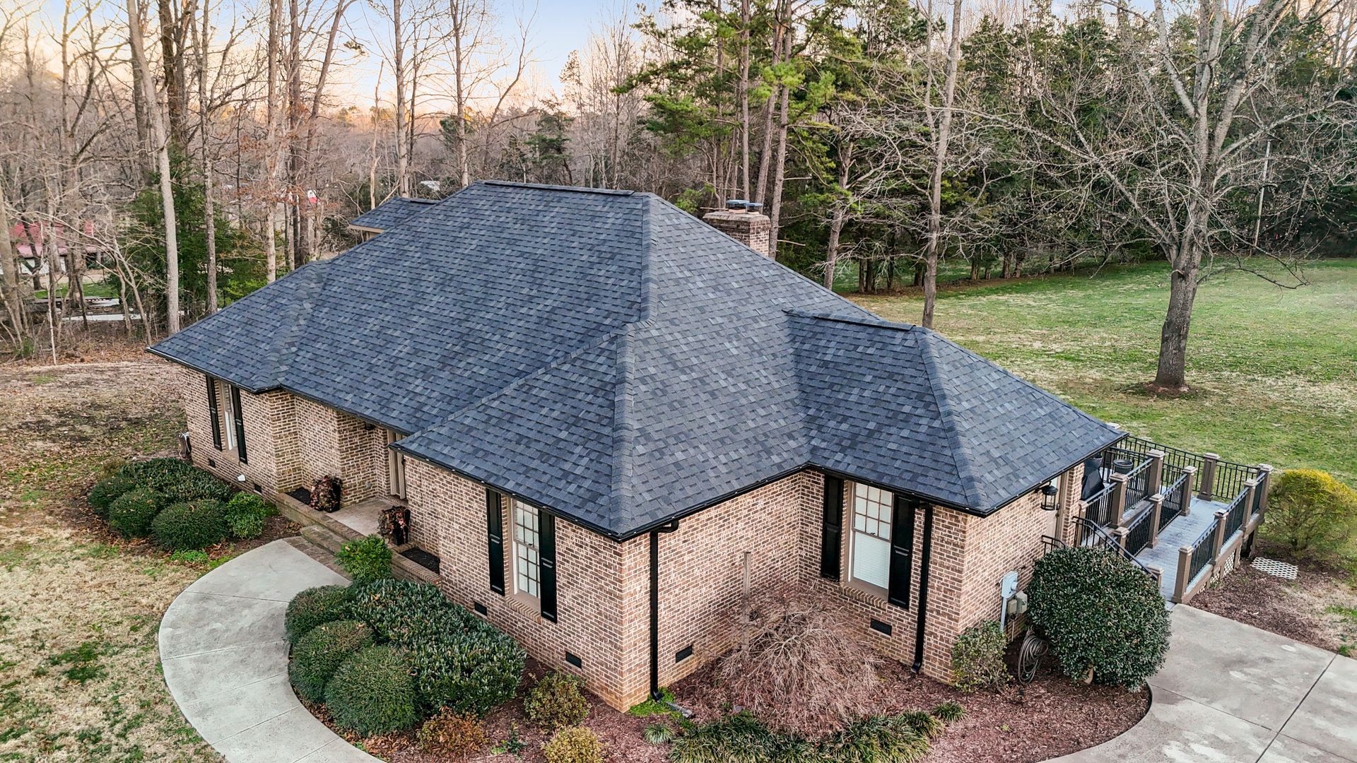 Brick house with dark roof, surrounded by trees and greenery; circular driveway.