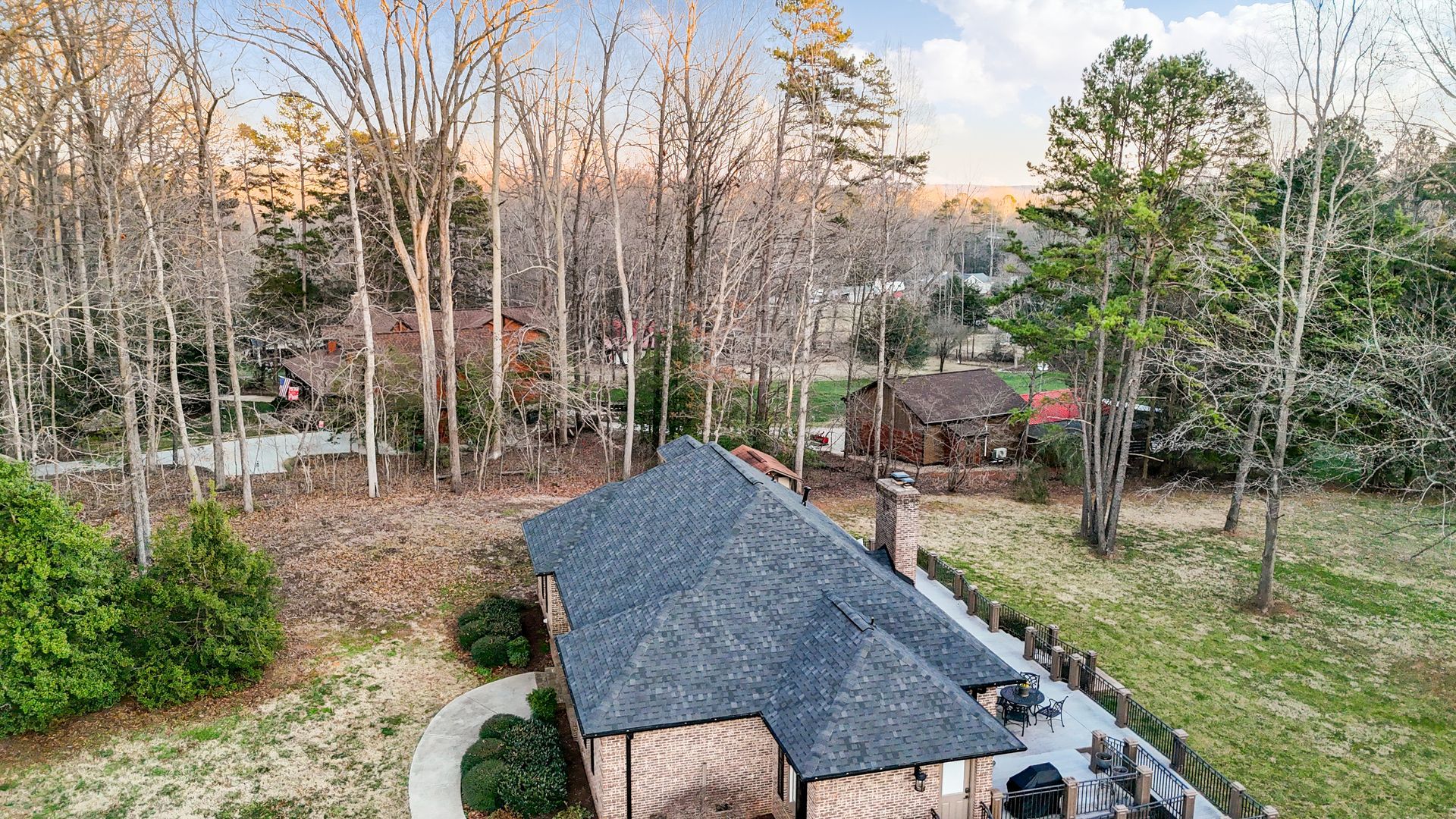 Aerial view of a dark-roofed house among trees in a grassy area with a glimpse of other homes in the distance.