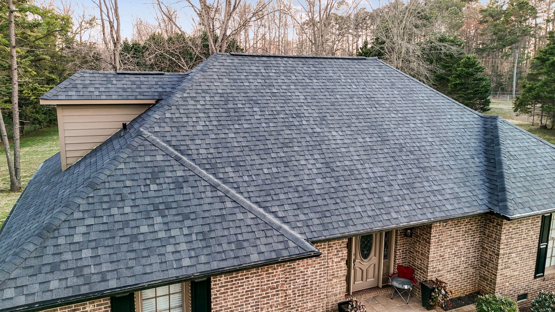 A house with a dark gray shingle roof and a brick exterior, set in a yard with trees.