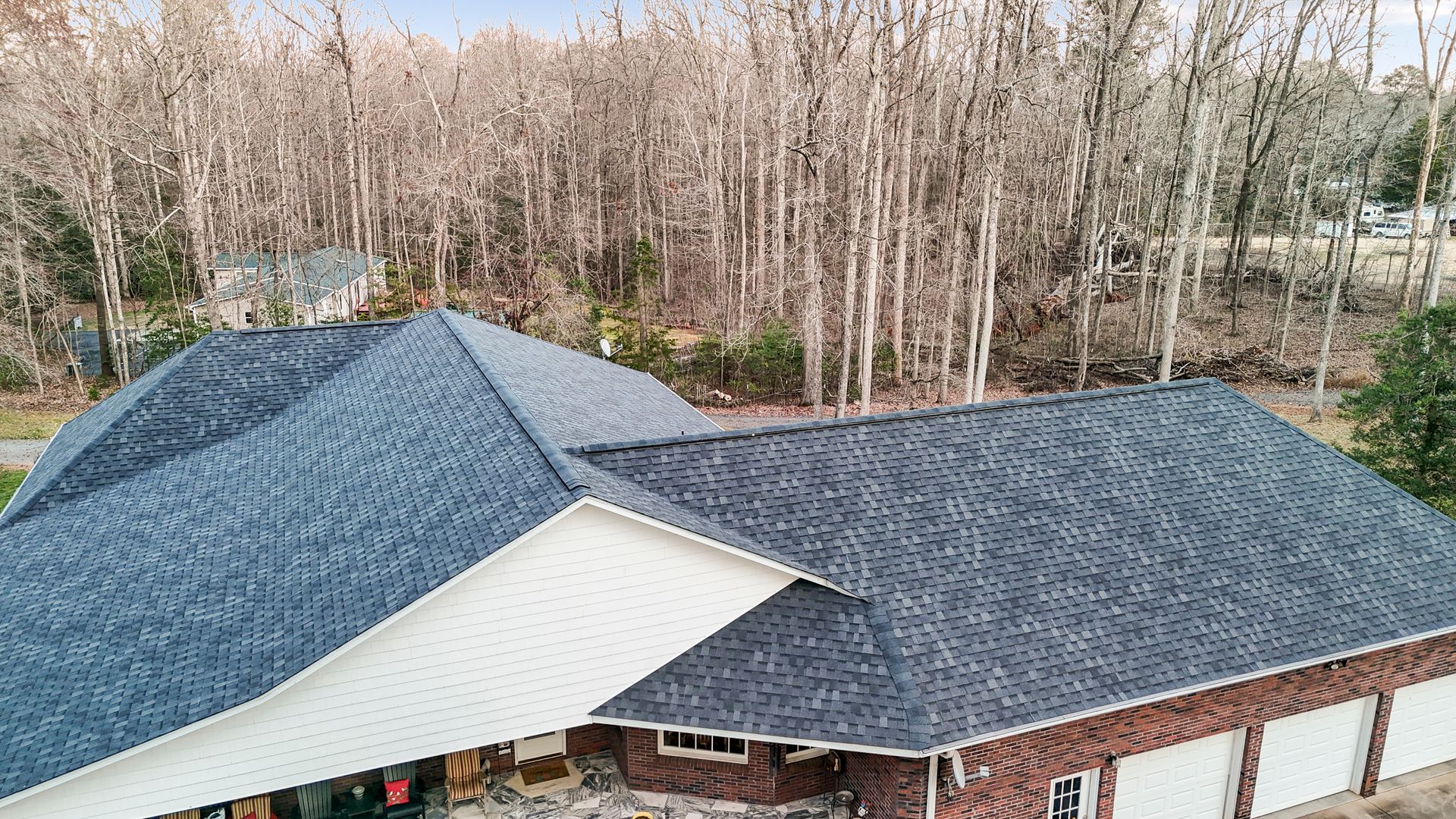 House with dark gray shingle roofs, brick and white siding, and a forest in the background.