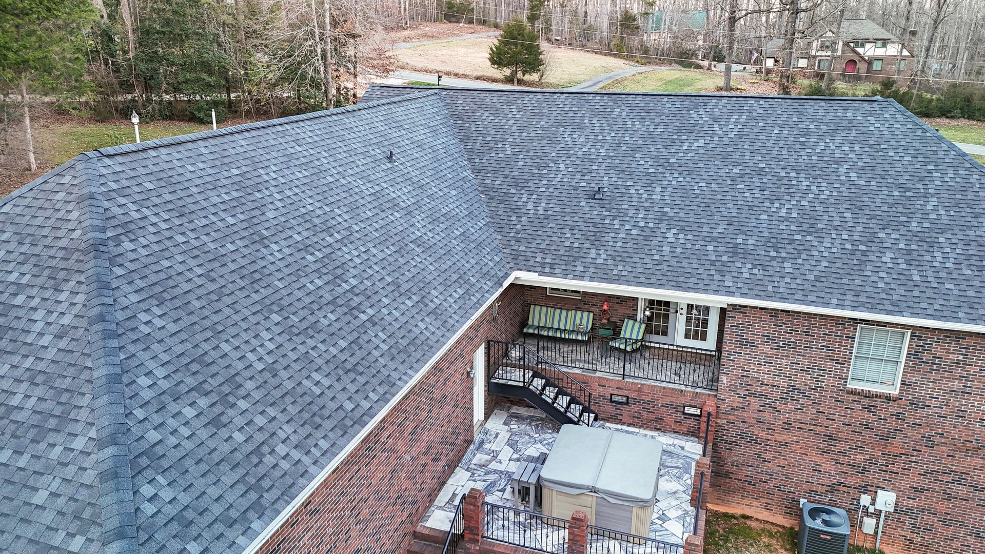 Dark gray shingled roof of a brick building, viewed from above, with a few trees in the background.