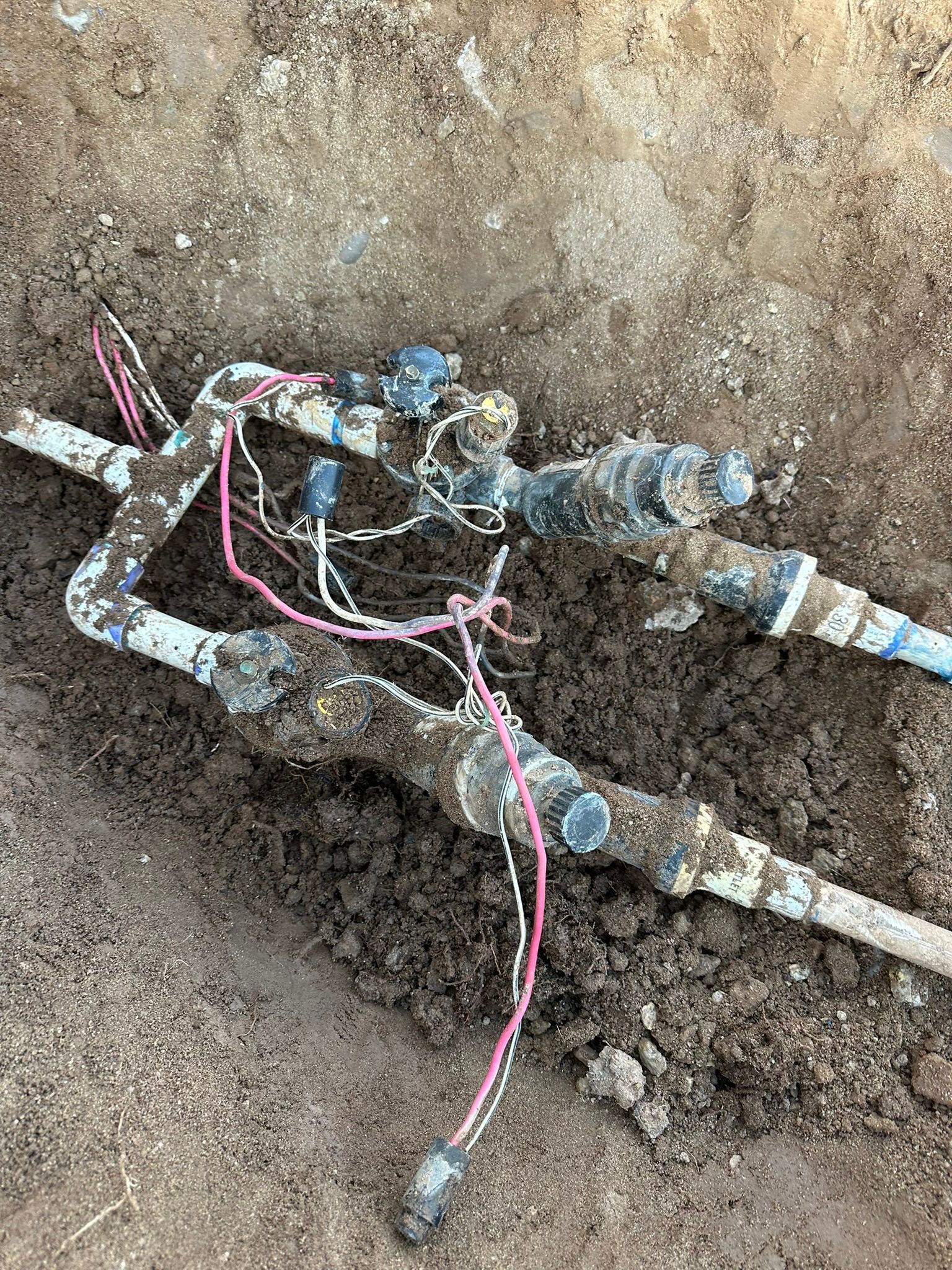 Irrigation system components, including pipes and valves with wiring, are exposed in a muddy trench.