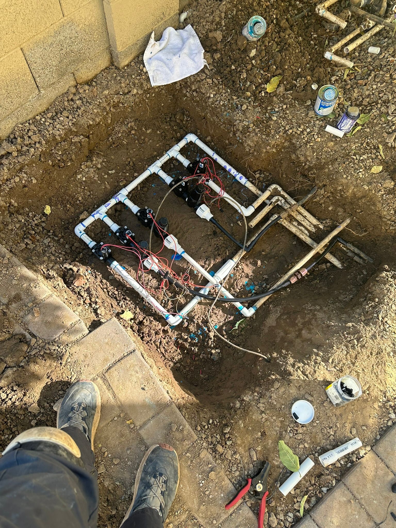 A person stands near exposed irrigation system pipes and wires in a dirt pit, amidst brick pavers.