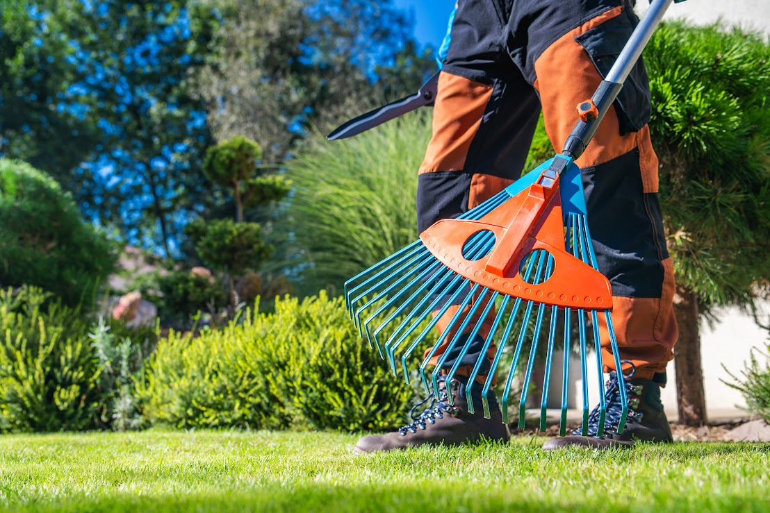 Gardener raking a manicured lawn, wearing orange work pants and boots. Green bushes and trees in the background.