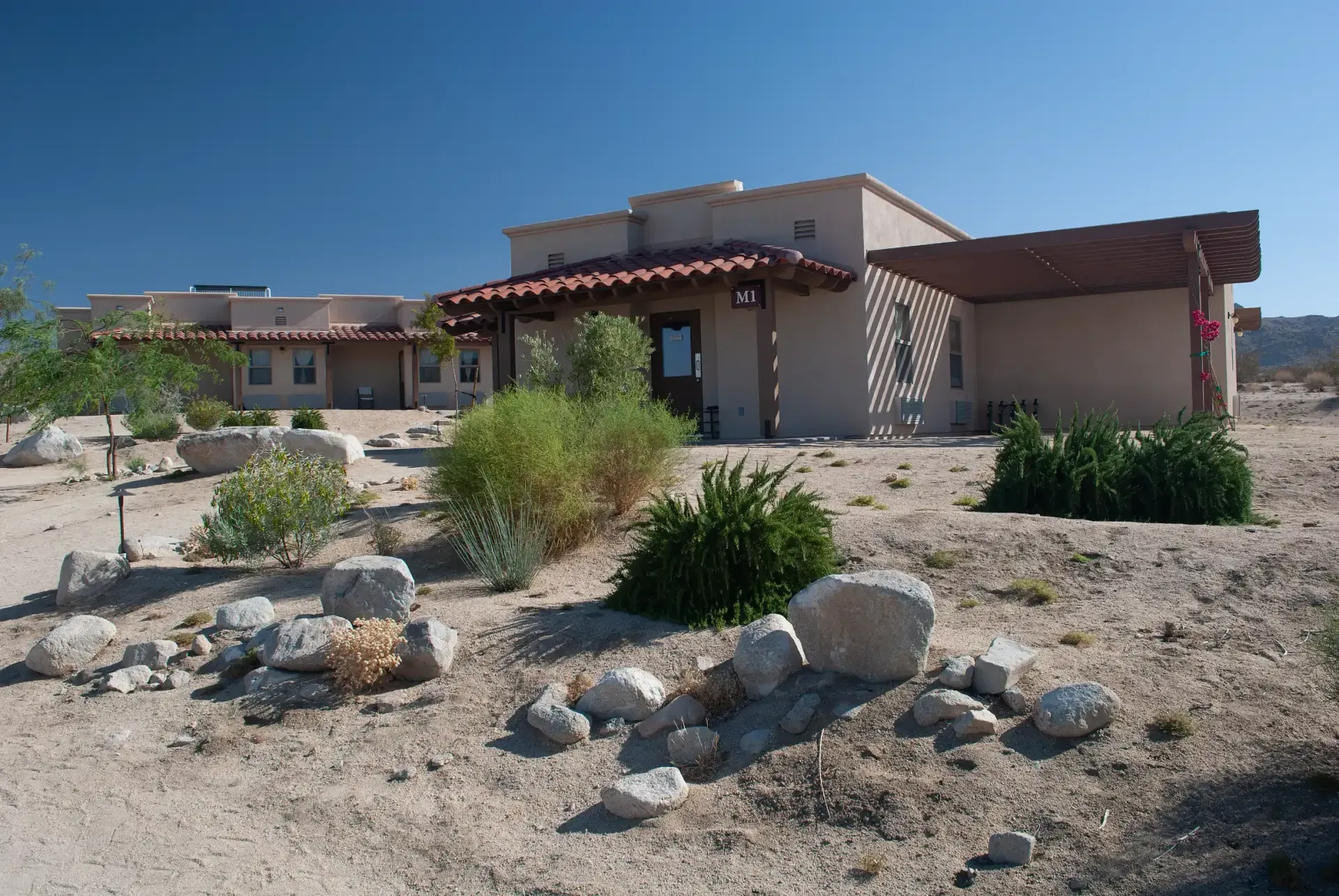 Adobe-style building in desert landscape with beige walls, red-tiled roof, and scattered rocks and shrubs in the foreground.