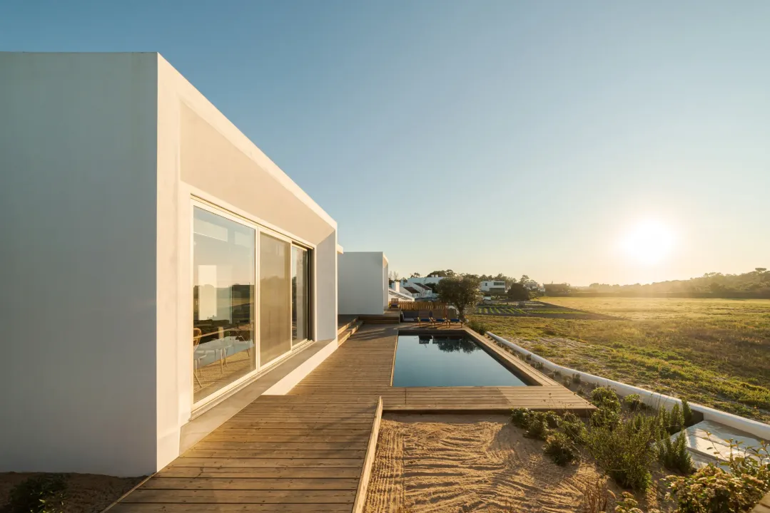 Modern white building with a wooden deck and rectangular pool, set against a field at sunset.