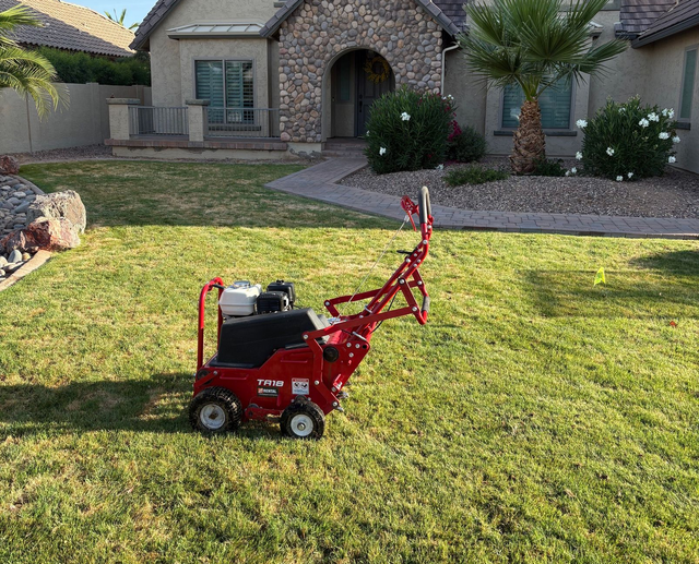 Man mowing a green lawn with a teal and black electric lawnmower. He wears a plaid shirt, jeans, gloves, and sneakers.