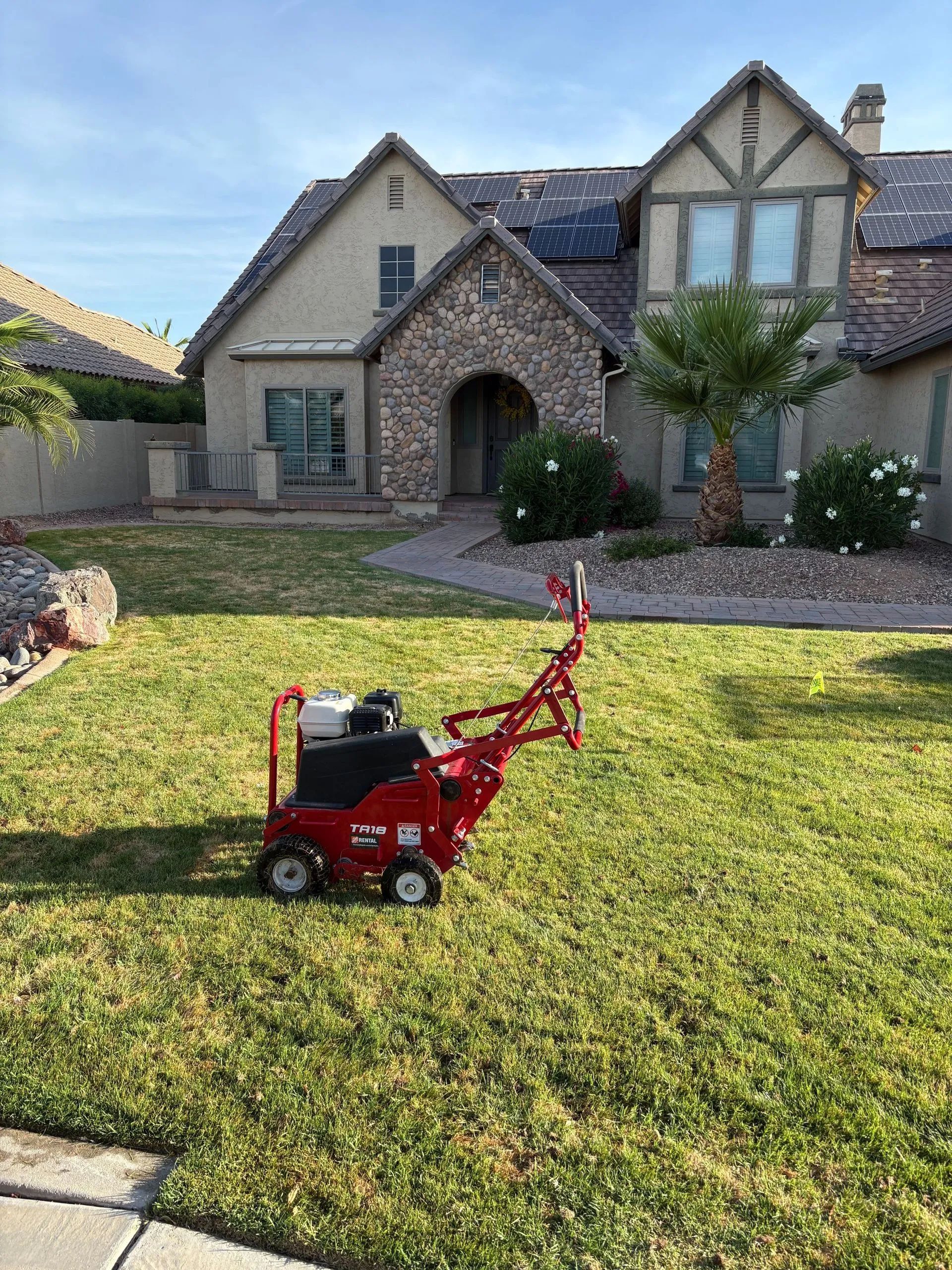 A red sod cutter sits on a green residential lawn in front of a tan, multi-story home with a stone facade.