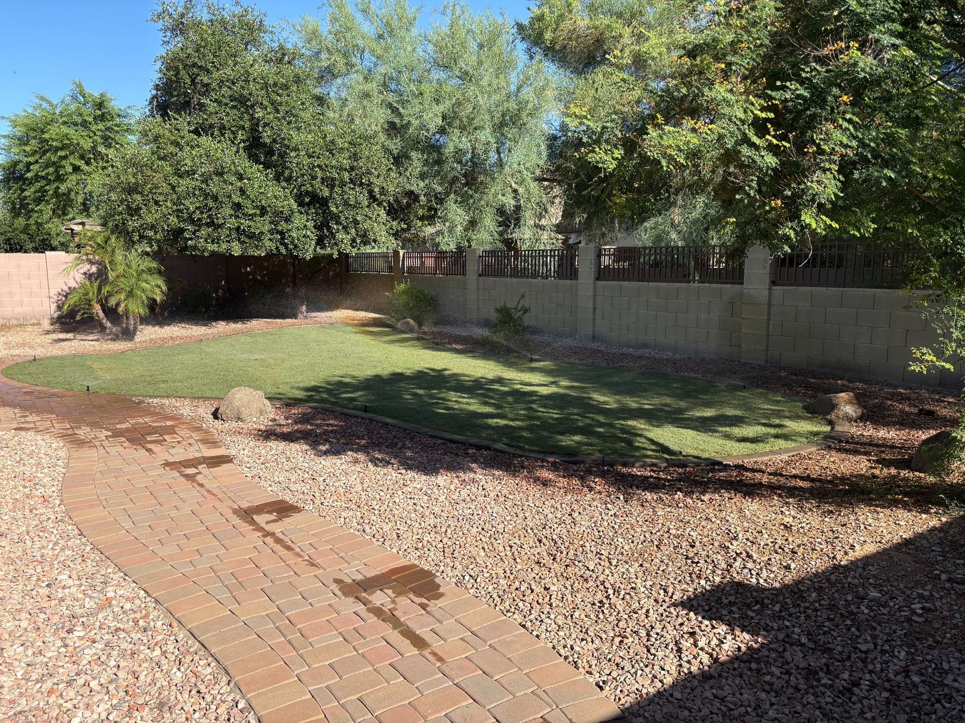 Backyard with a brick path, grass, trees, and a gravel area under a bright sky.