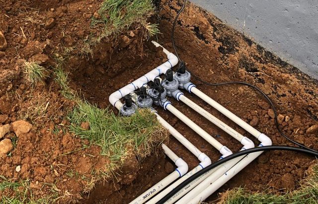 Irrigation system manifold with multiple valves and PVC pipes in a trench, near grass and a paved surface.