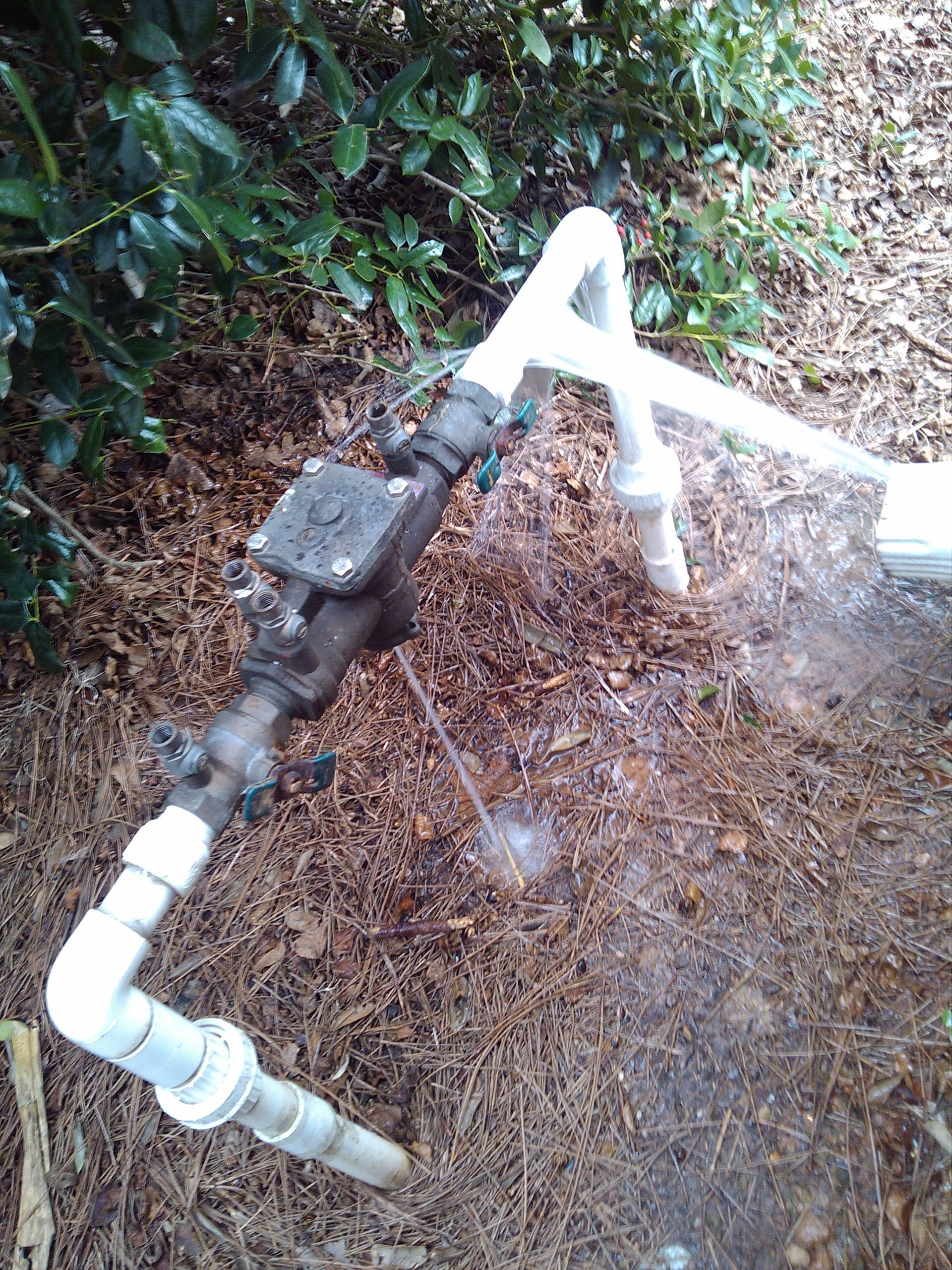 A backflow preventer system with white PVC pipes, metal valve, and dark earth setting against green foliage.