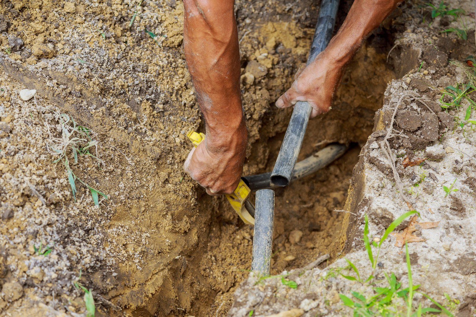 Hands cutting a black pipe in a dirt trench. The person is using a yellow tool, likely for plumbing