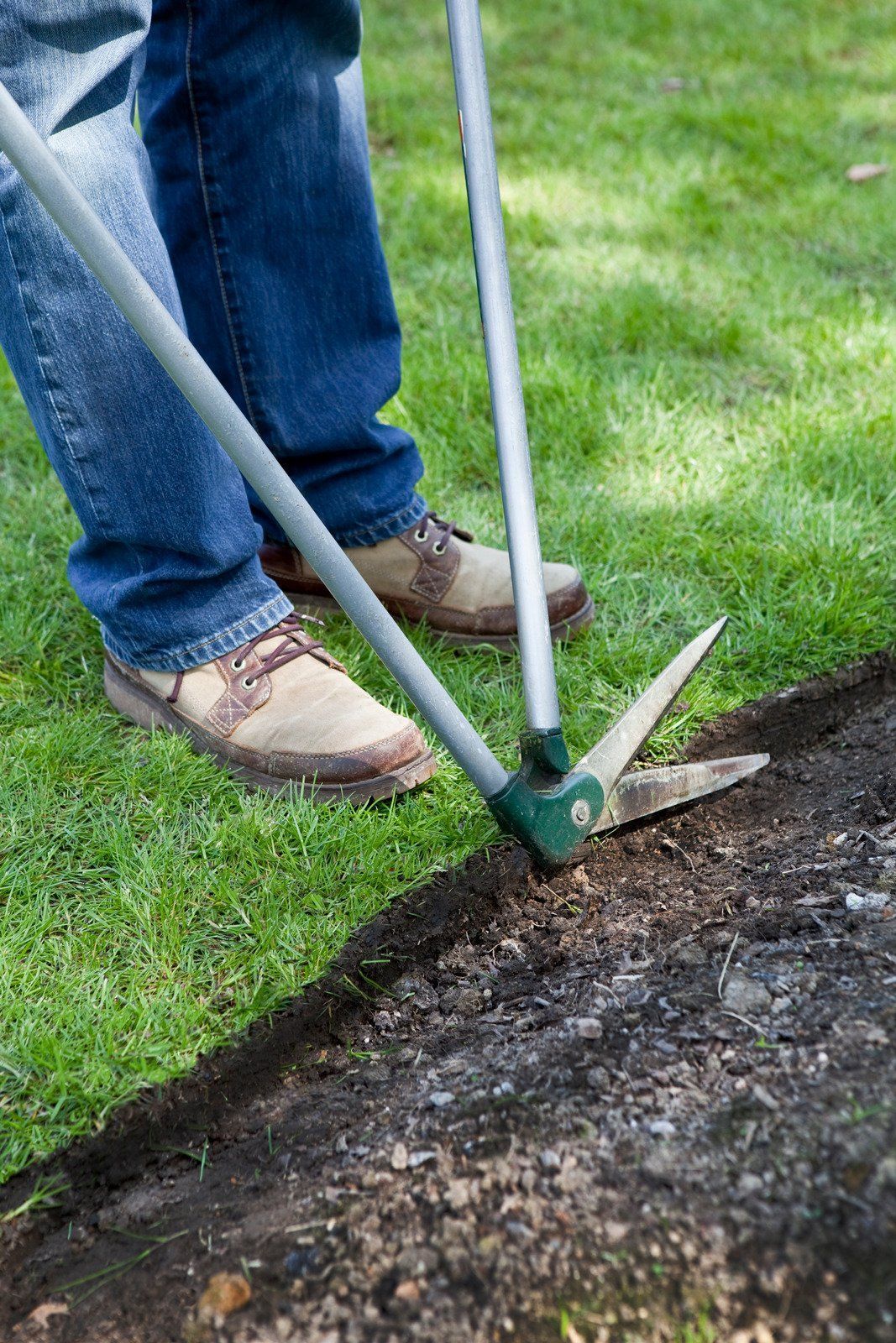 Person edging a lawn with long-handled shears, trimming the edge of a dark, gravelly area next to green grass.