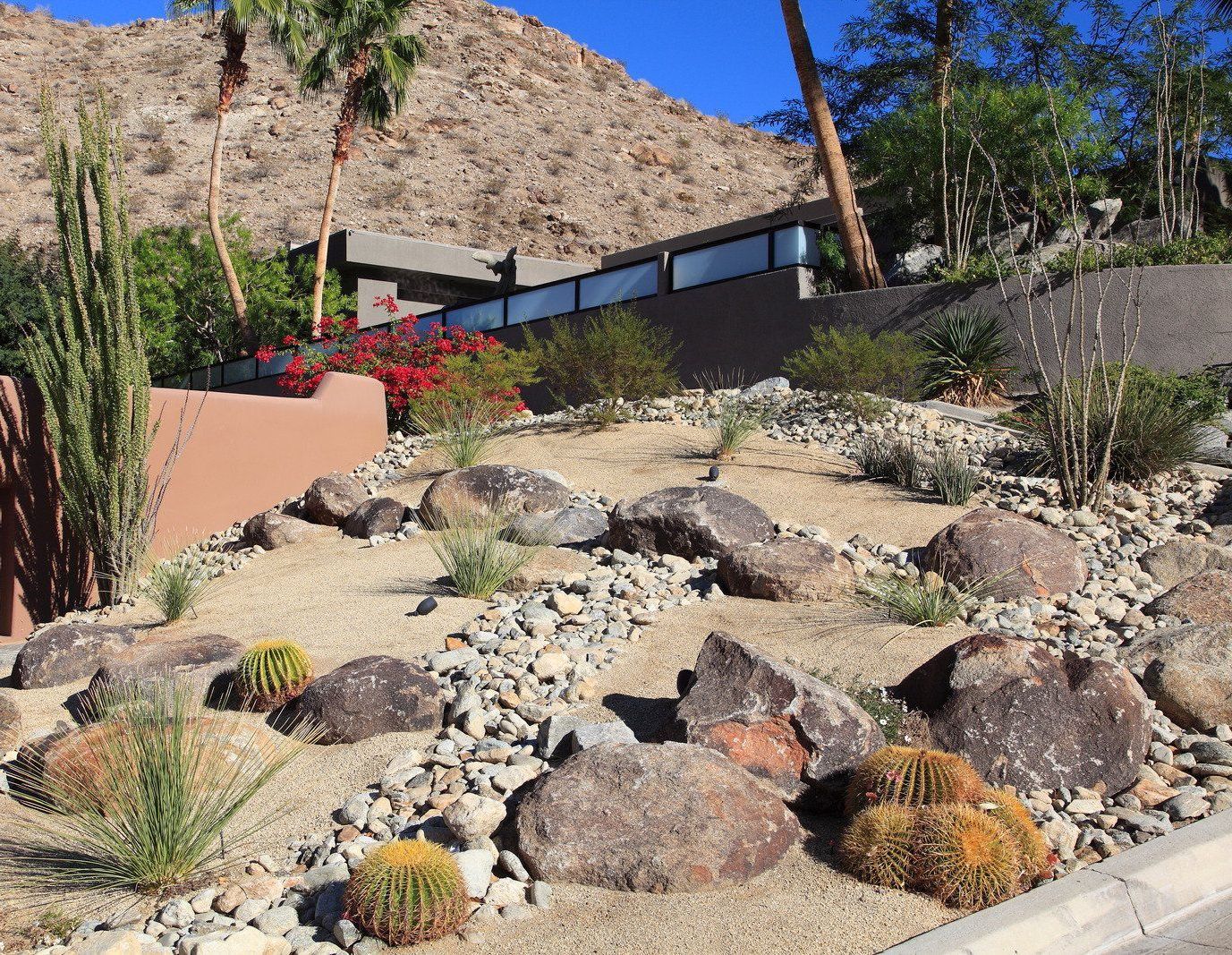 Desert landscape with cacti, rocks, and a building against a mountain backdrop under a blue sky.
