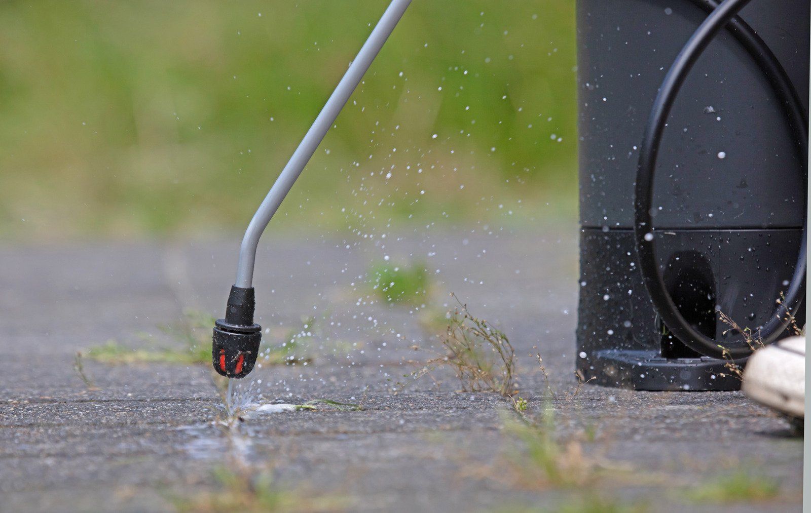 A sprayer nozzle releasing liquid onto a paved surface with grass nearby, next to a black container.
