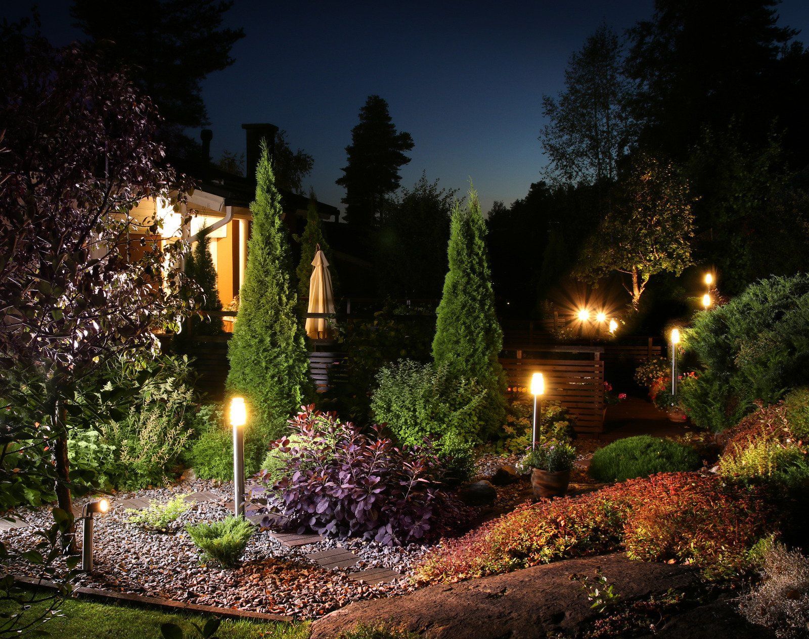 Nighttime garden with illuminated pathways, trees, and landscaping in front of a house with lights on.