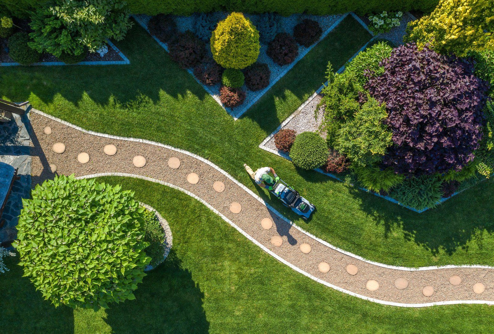 Overhead view of a landscaped garden with a winding stone path, various shrubs, and a person mowing the lawn.