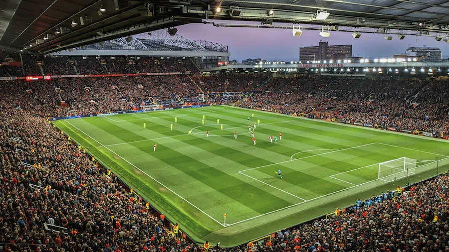 Soccer match in a packed stadium; green field, players, cheering crowd, bright lights at dusk.