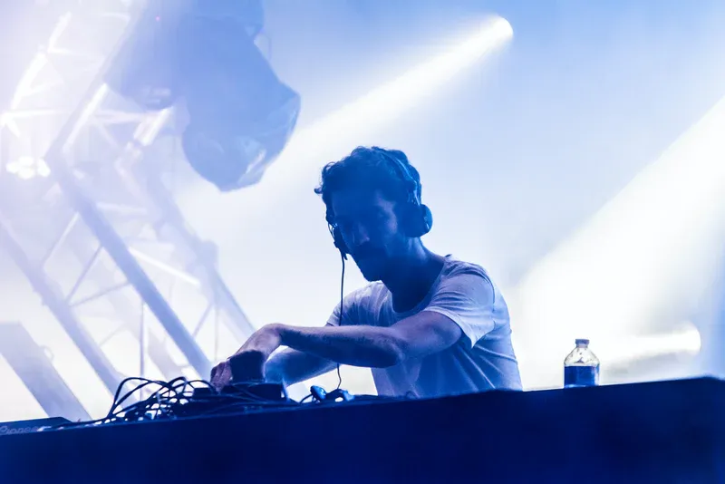 DJ performing at a concert, bathed in blue light. He is wearing headphones and operating music equipment.