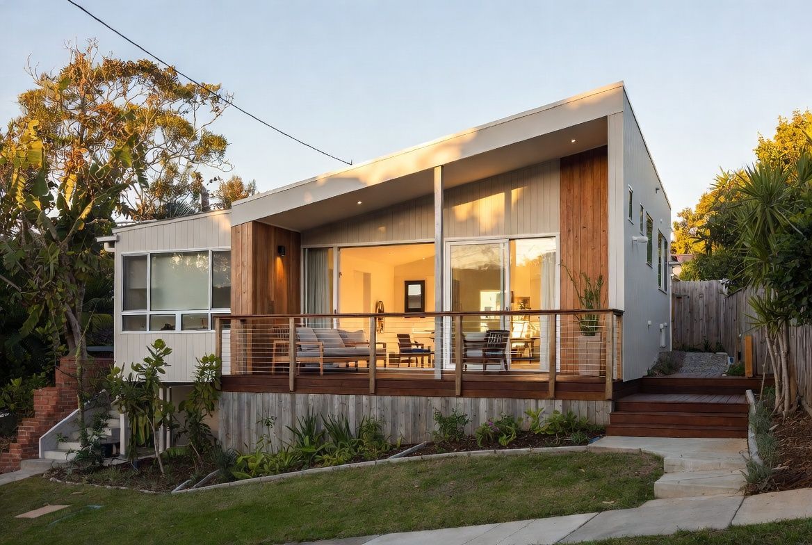 Modern house with large windows and a wooden porch at sunset, surrounded by trees and a green lawn