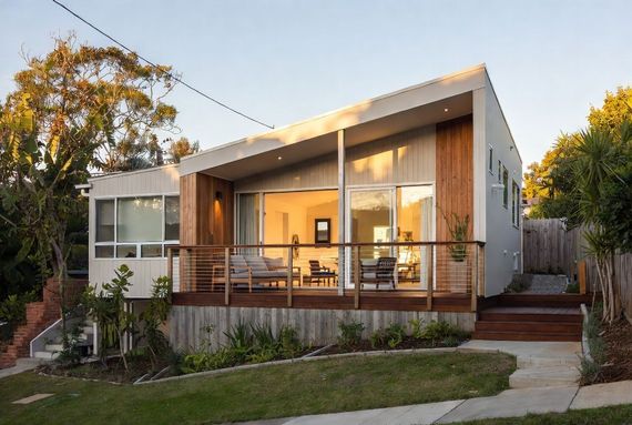 Modern house with large windows and a wooden porch at sunset, surrounded by trees and a green lawn