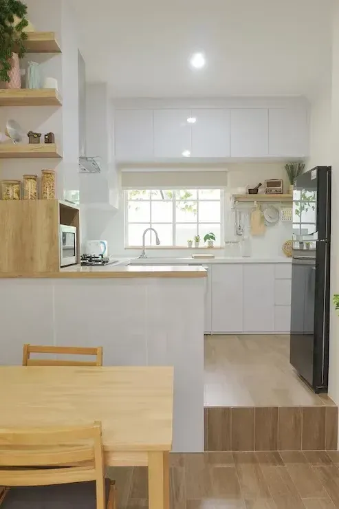 Bright minimalist kitchen and dining area with light wood table, white cabinets, and window over the sink