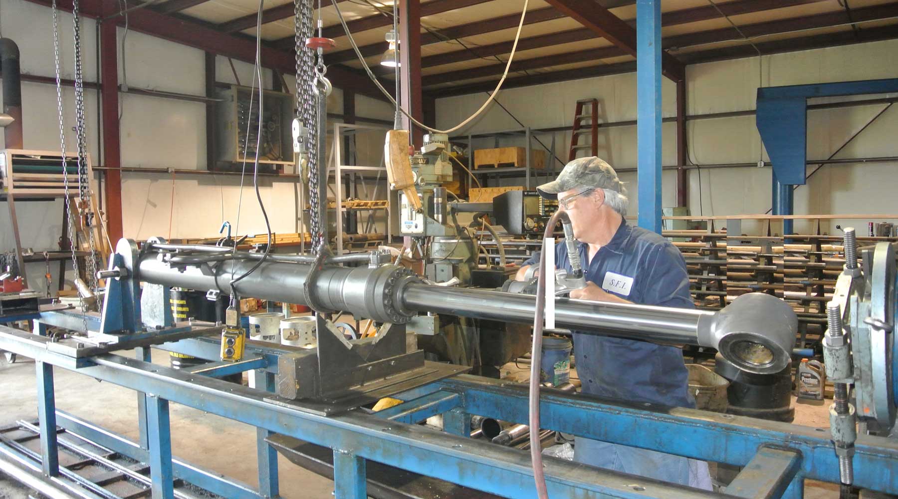 Man working on a metal cylinder in a workshop, using machinery, under a hanging chain.
