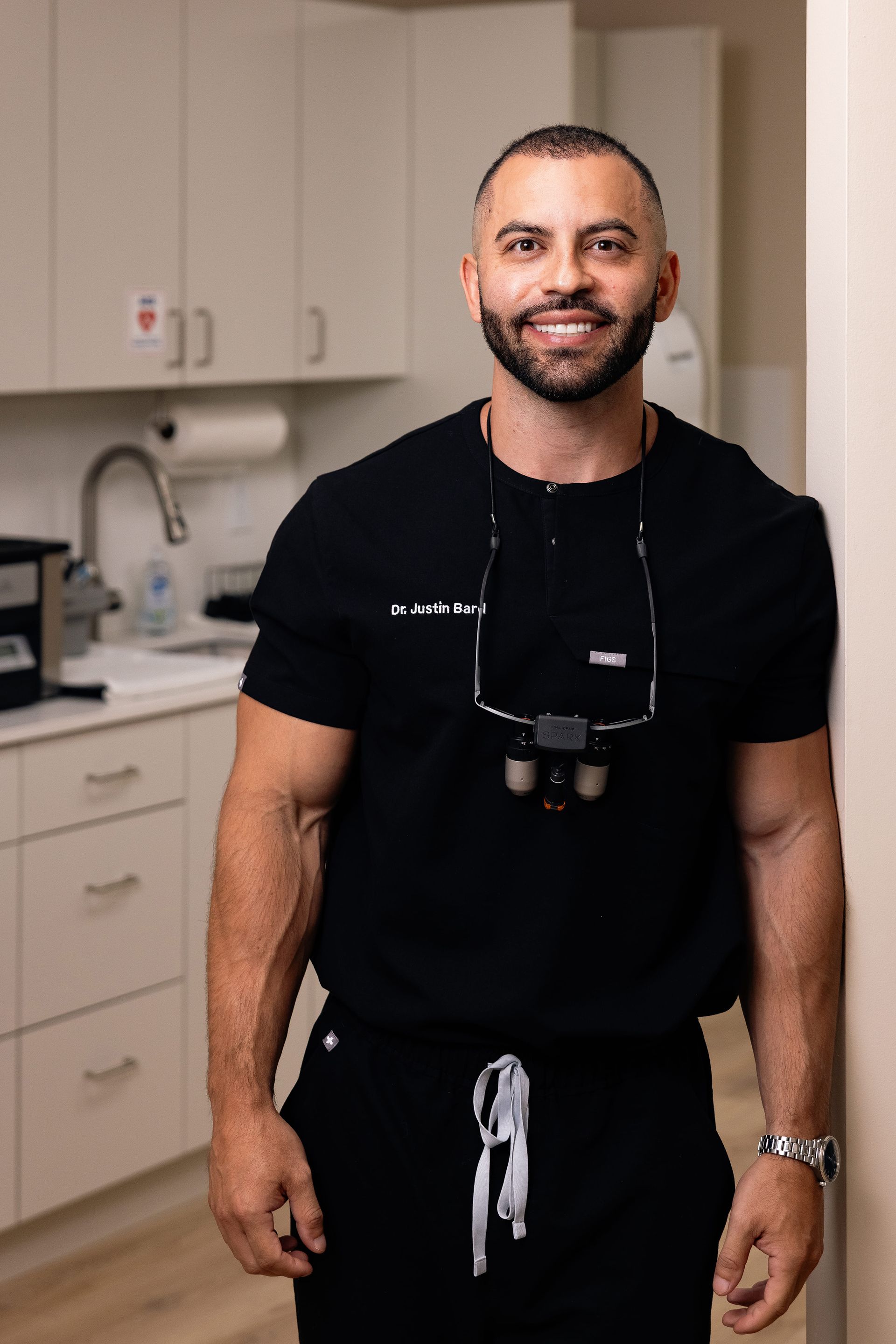 Man in black scrubs smiles, leaning against wall in dental office.