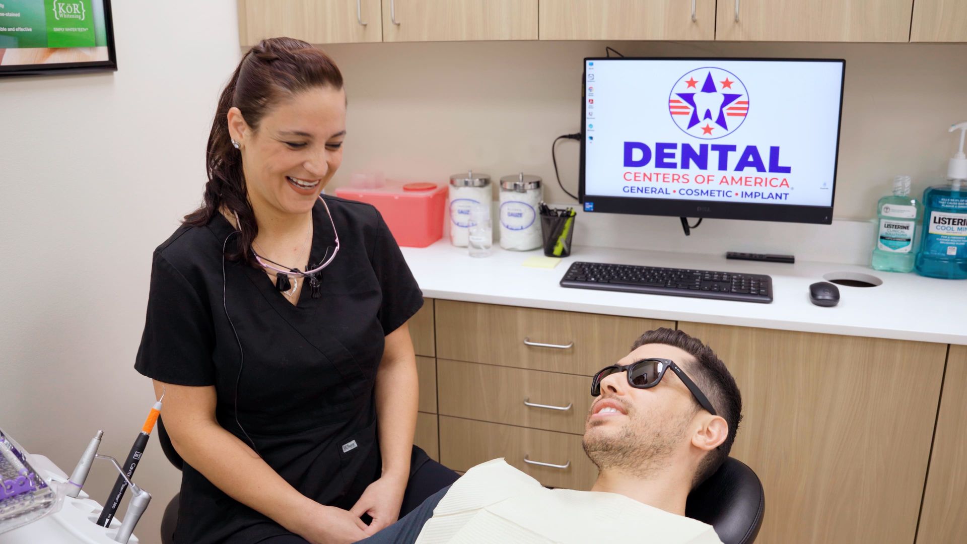 Dental assistant smiles at patient in a dental chair, computer in the background.