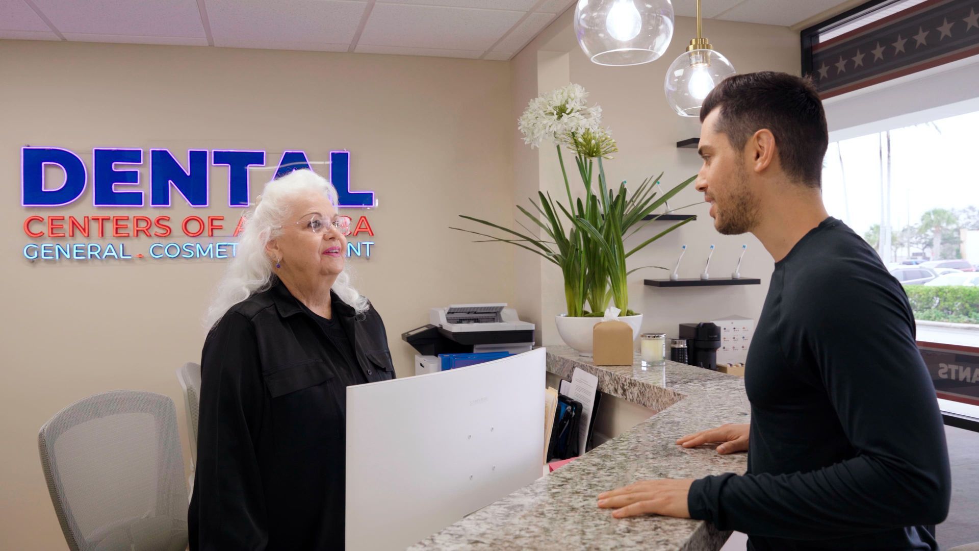 Man at dental office reception desk talks to a woman with white hair; 