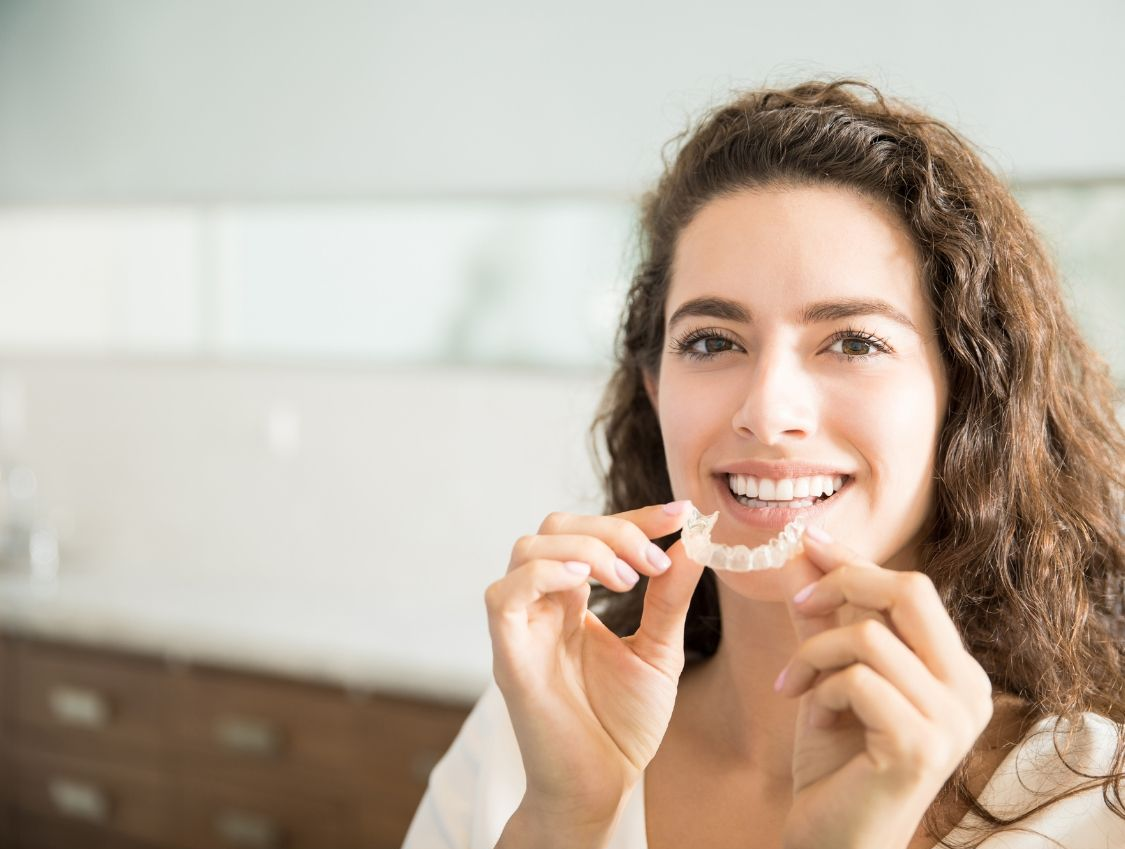 Woman smiling, holding a clear aligner, likely for teeth straightening in a bright room.