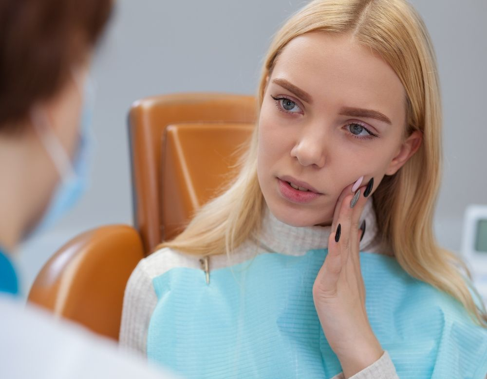 Blonde woman in dental chair, touching cheek, appears to have toothache. Doctor with mask consults.