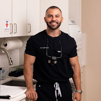 Man in black scrubs smiles in a dental office. He is muscular, wearing glasses, and a watch.