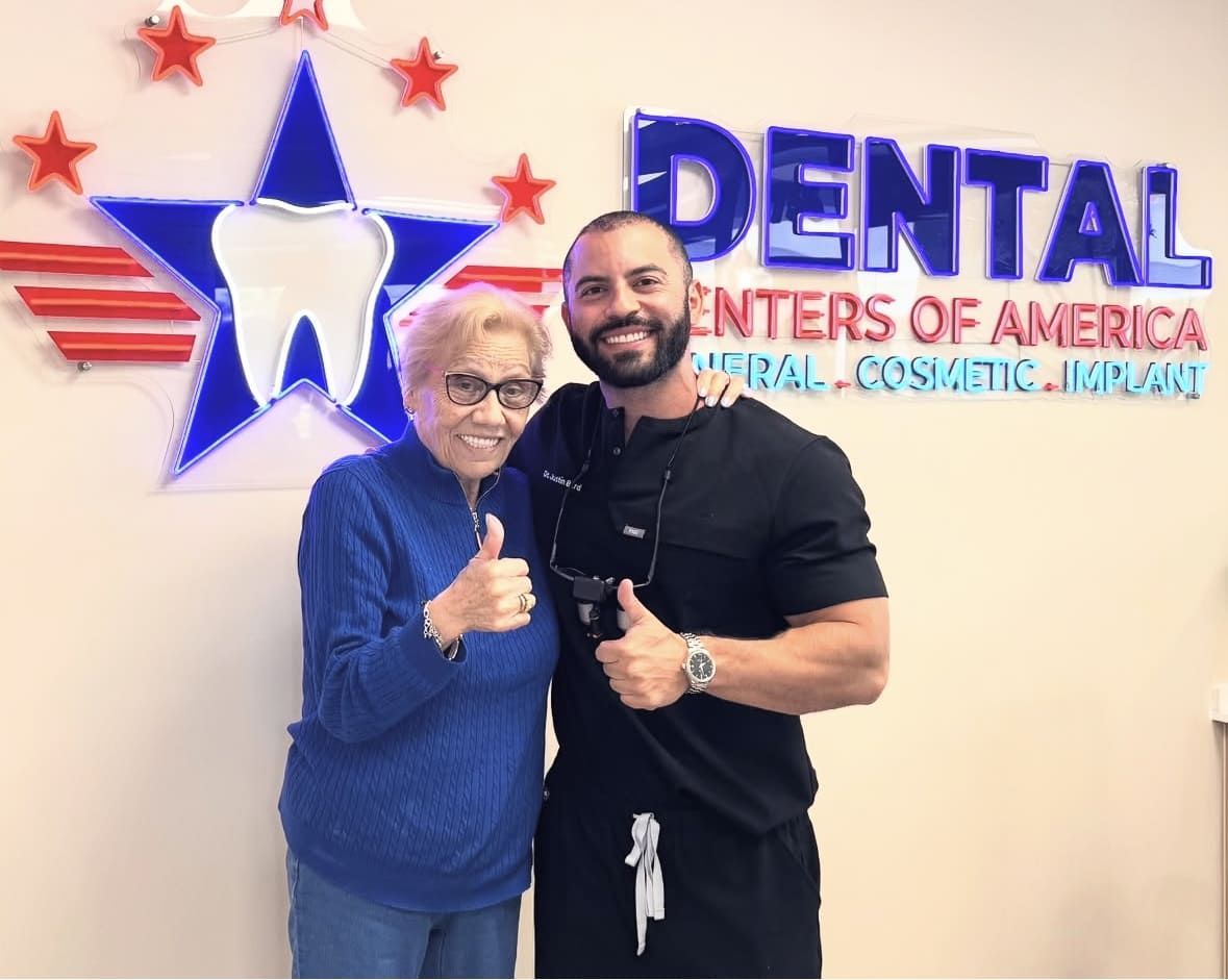 Dentist and elderly patient smiling, giving thumbs up in front of a Dental Centers of America sign.