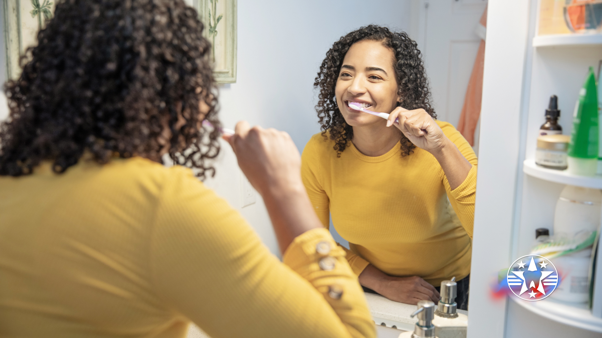 Woman brushing teeth in bathroom, reflected in mirror. She wears yellow and smiles.
