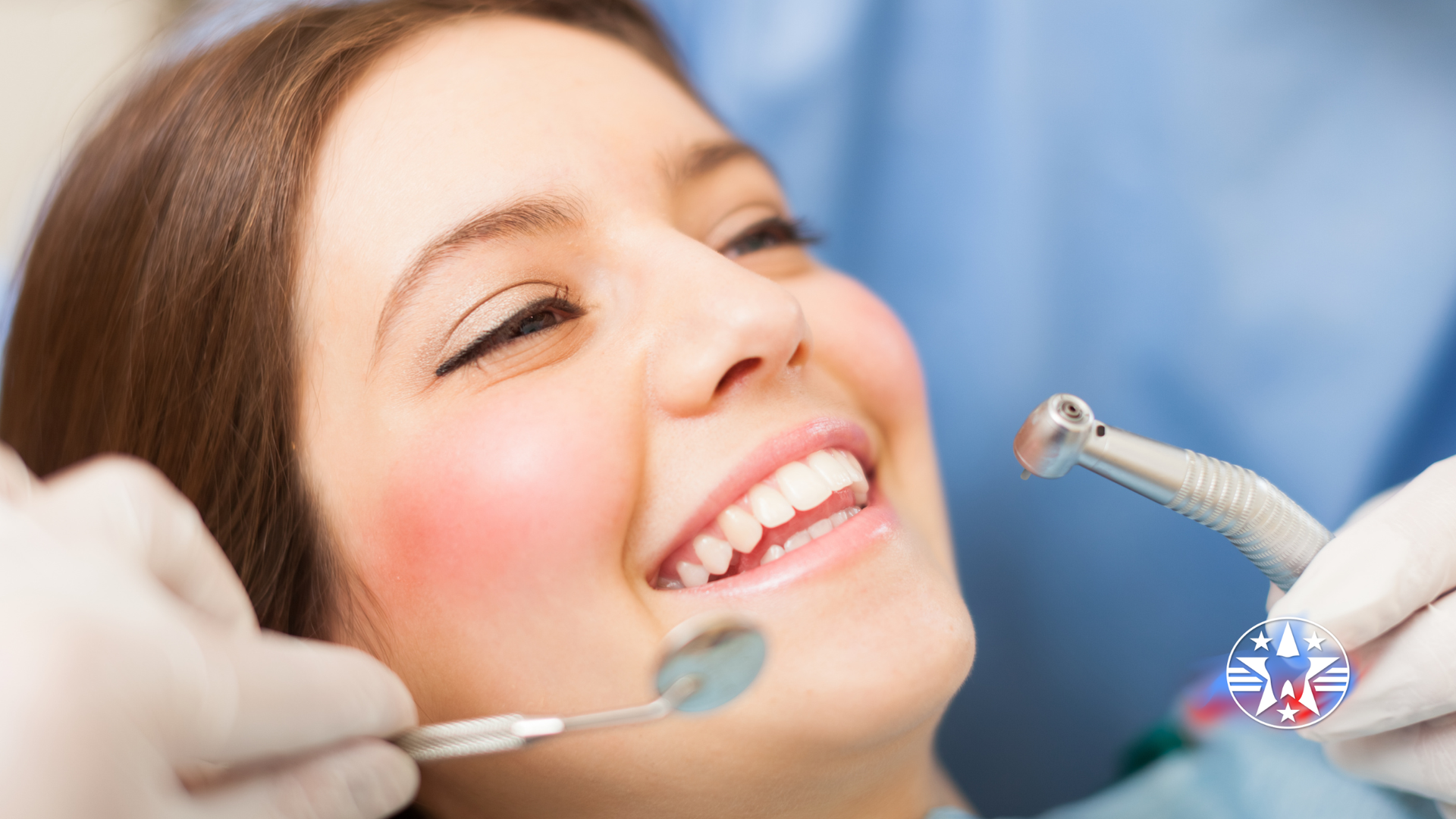 Woman smiling during a dental exam; dentist holds tools, bright interior.