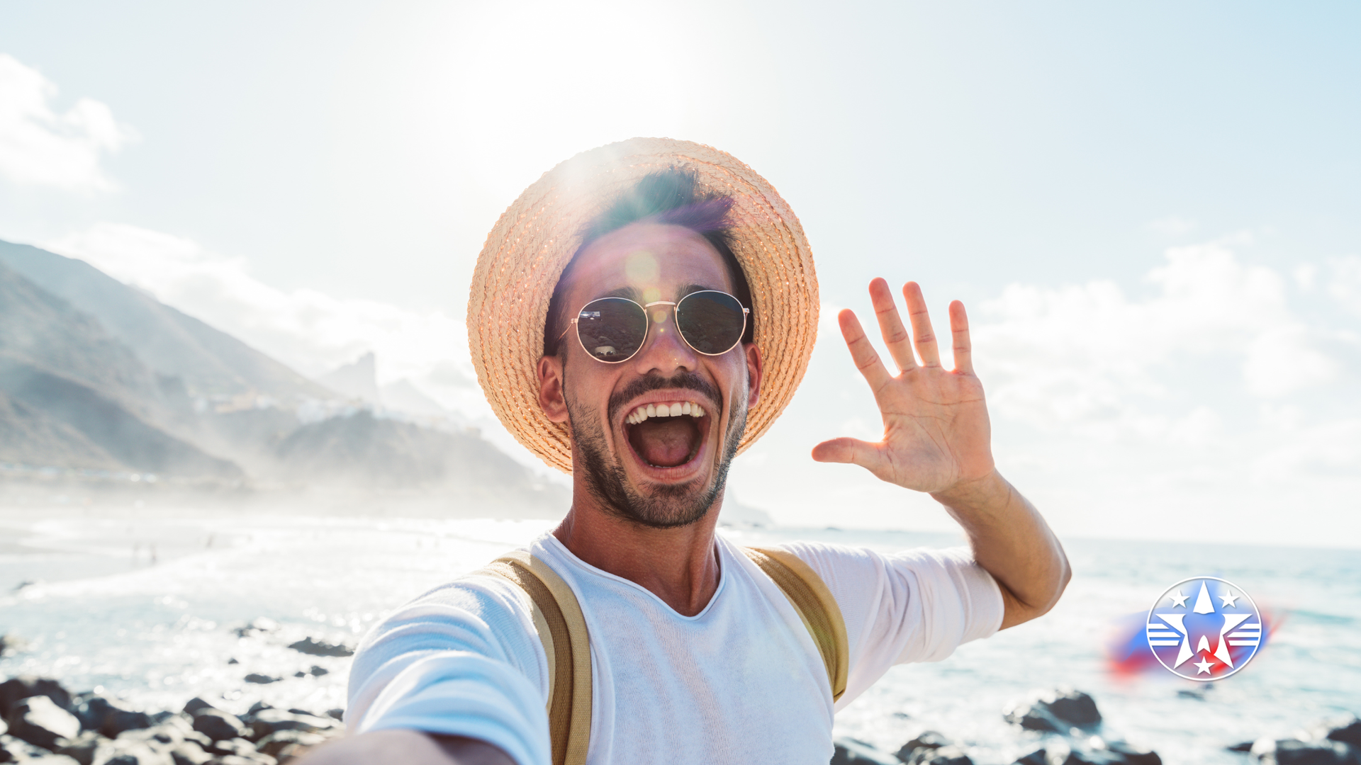 Man in straw hat and sunglasses waves with ocean and mountains in background.
