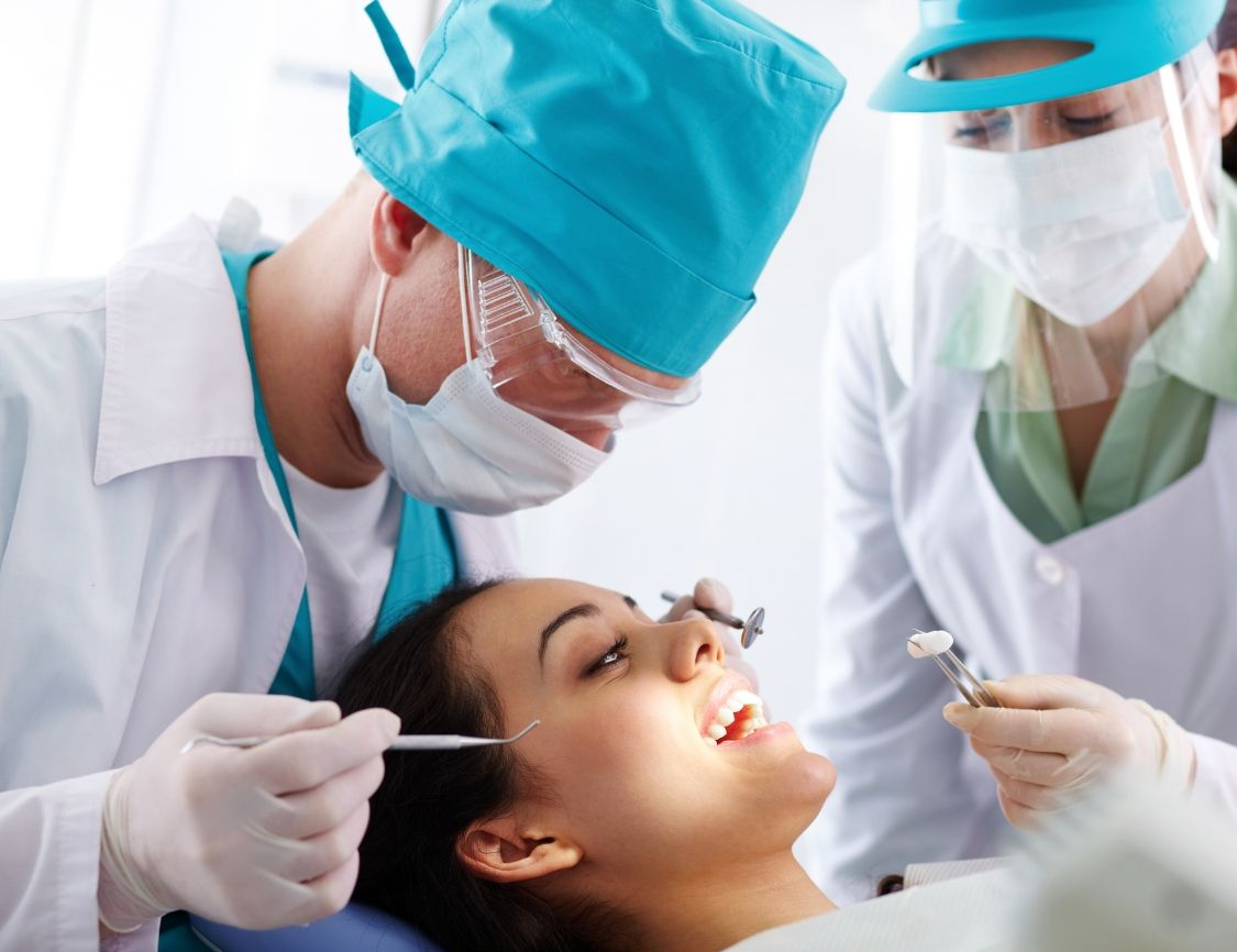 Dentist examining a patient's teeth; dental assistant present in a brightly lit office.