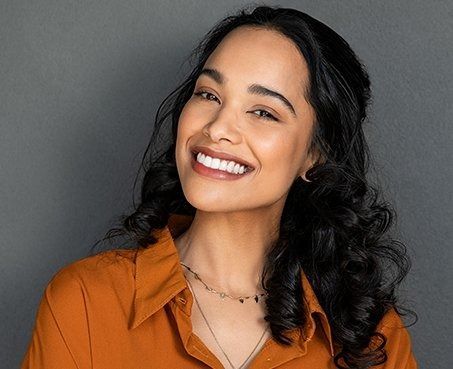 Woman with dark curly hair smiles, wearing orange shirt and necklace, against a gray background.
