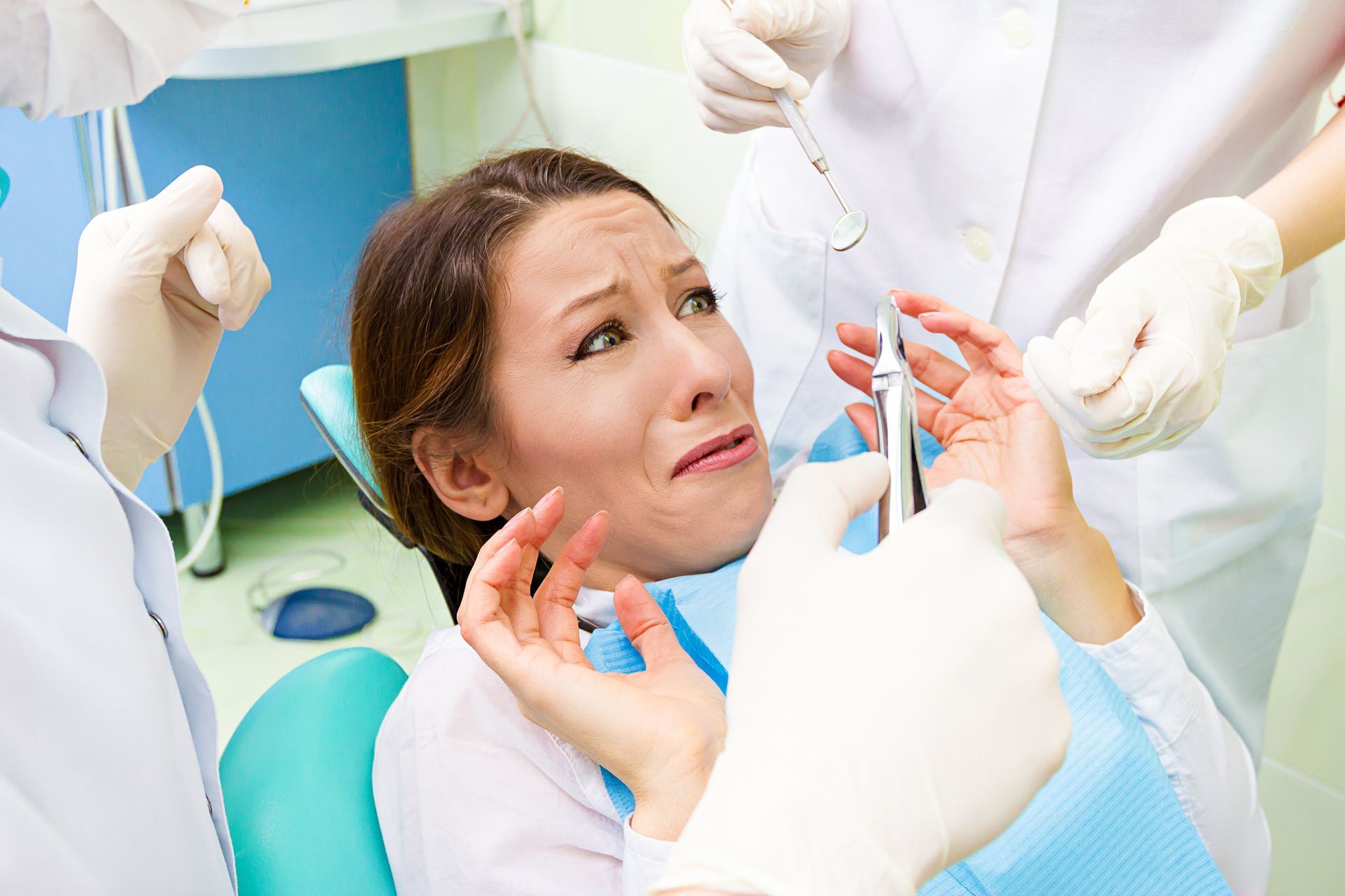 Woman in dentist chair, fearful expression, surrounded by gloved hands holding dental tools.