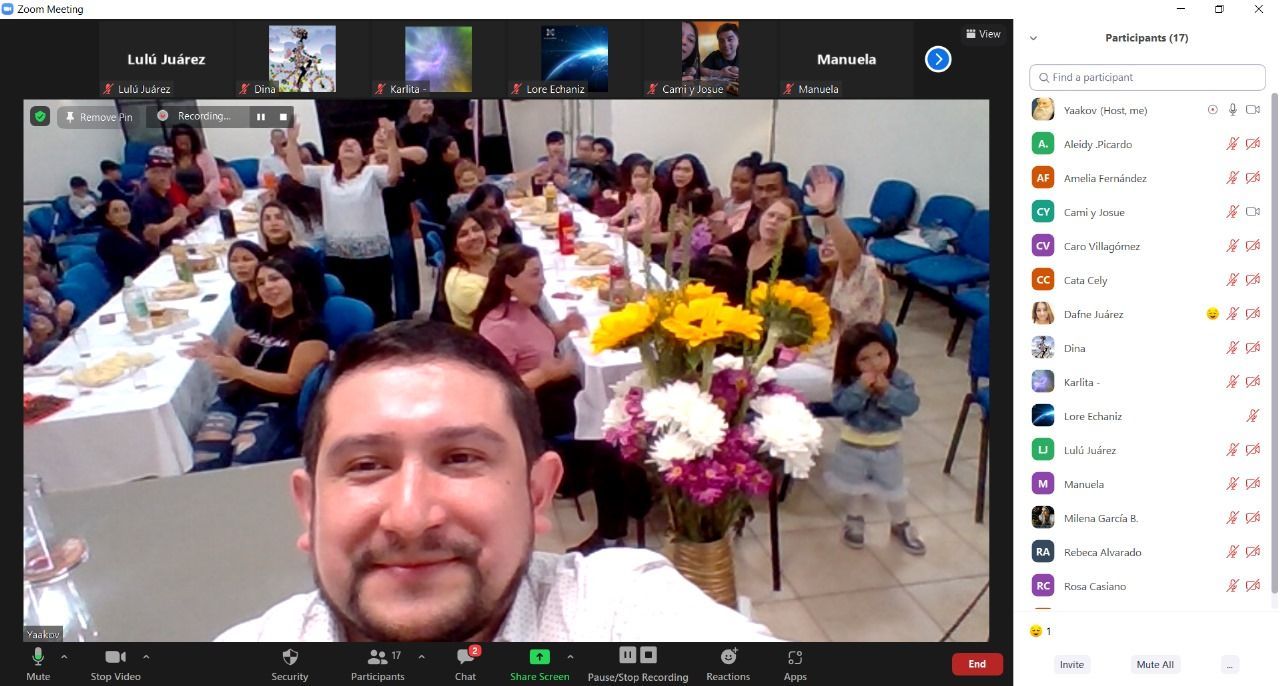 Un hombre sonríe durante una llamada de Zoom con un grupo sentado en una mesa larga, celebrando, con flores.
