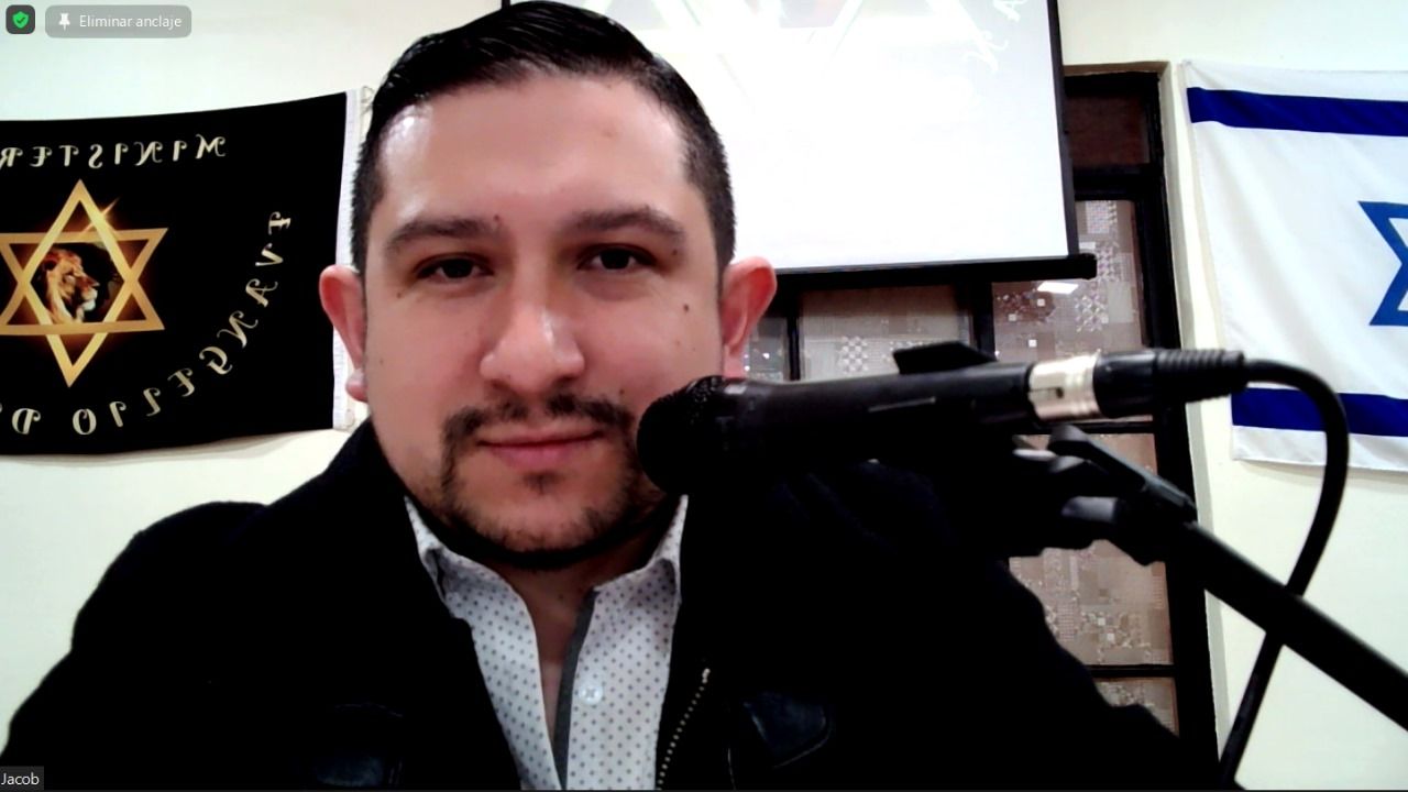 Un hombre con un micrófono sonríe, banderas con símbolos judíos visibles en el fondo.