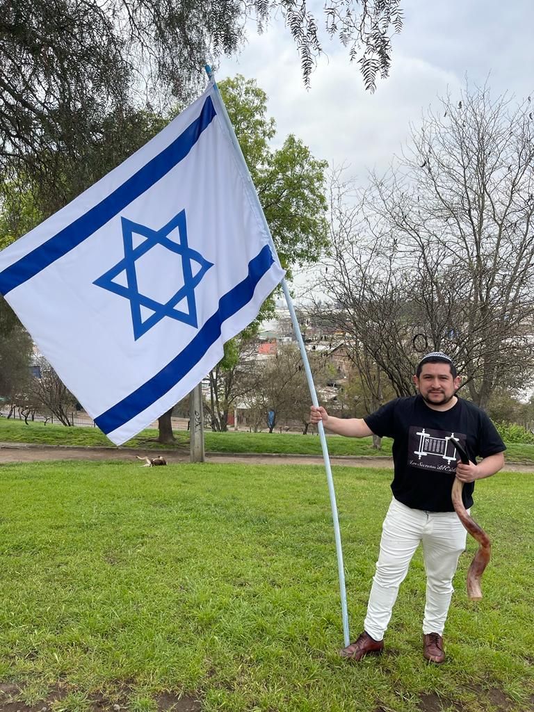 Hombre con bandera israelí al aire libre. Lleva camisa negra, pantalón blanco y zapatos marrones. Cielo nublado y vegetación.