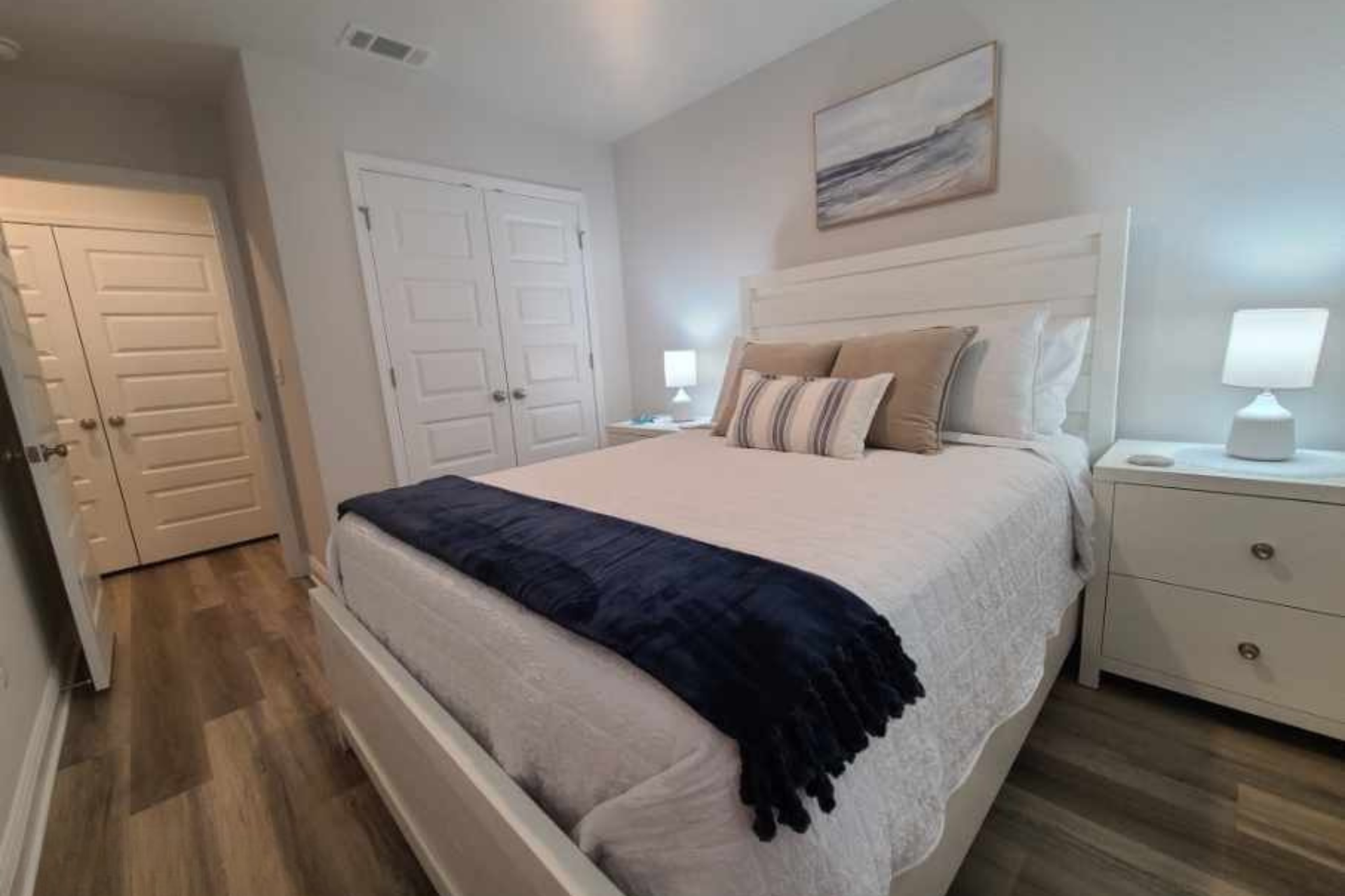 Bedroom with white bed, nightstands, and closed closet doors. Light walls and flooring.